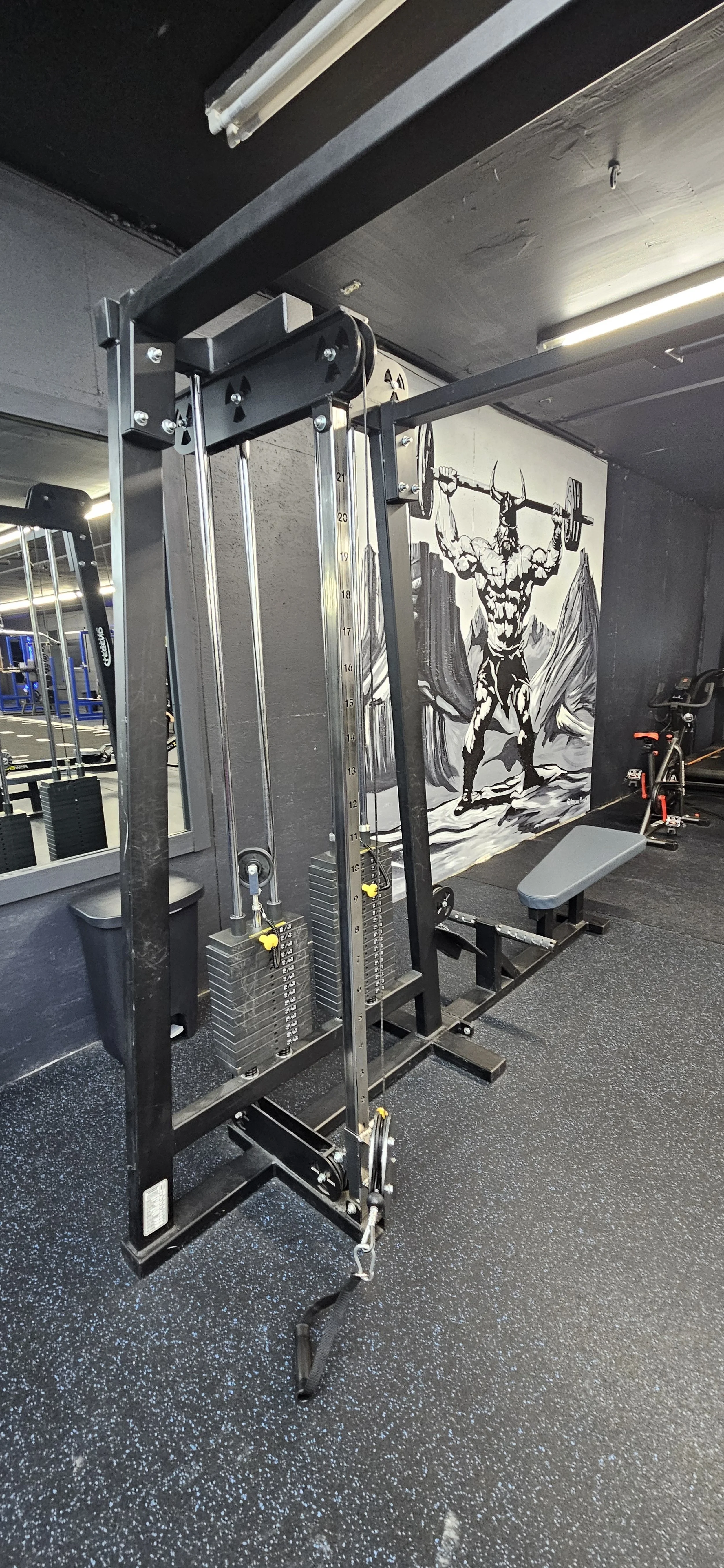 Black fitness machine equipment in a gym with a mural of a muscular figure holding a barbell against a mountain backdrop painted on the wall.