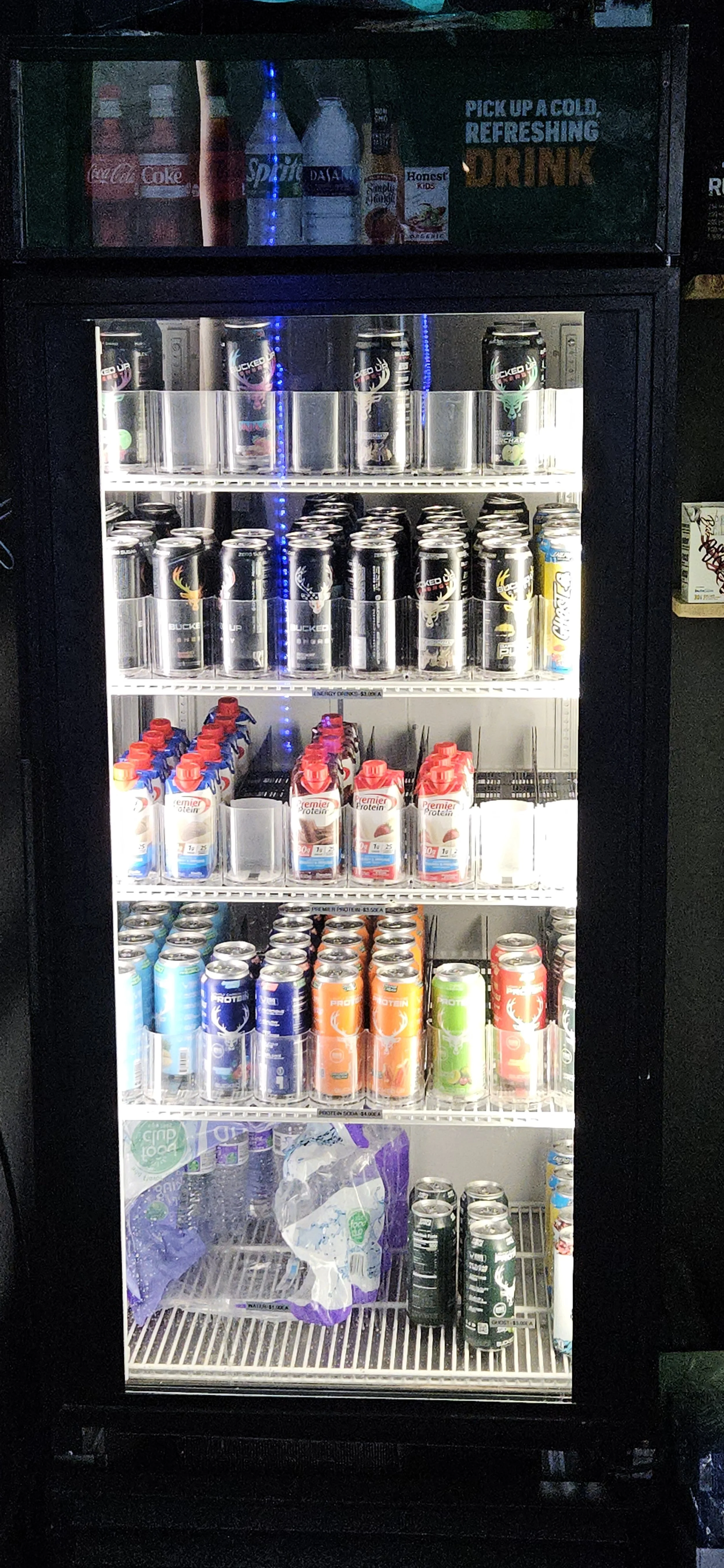 A refrigerated display case filled with various cans and bottles of beverages, including energy drinks, protein drinks, and soda, with some water bottles on the top shelf.