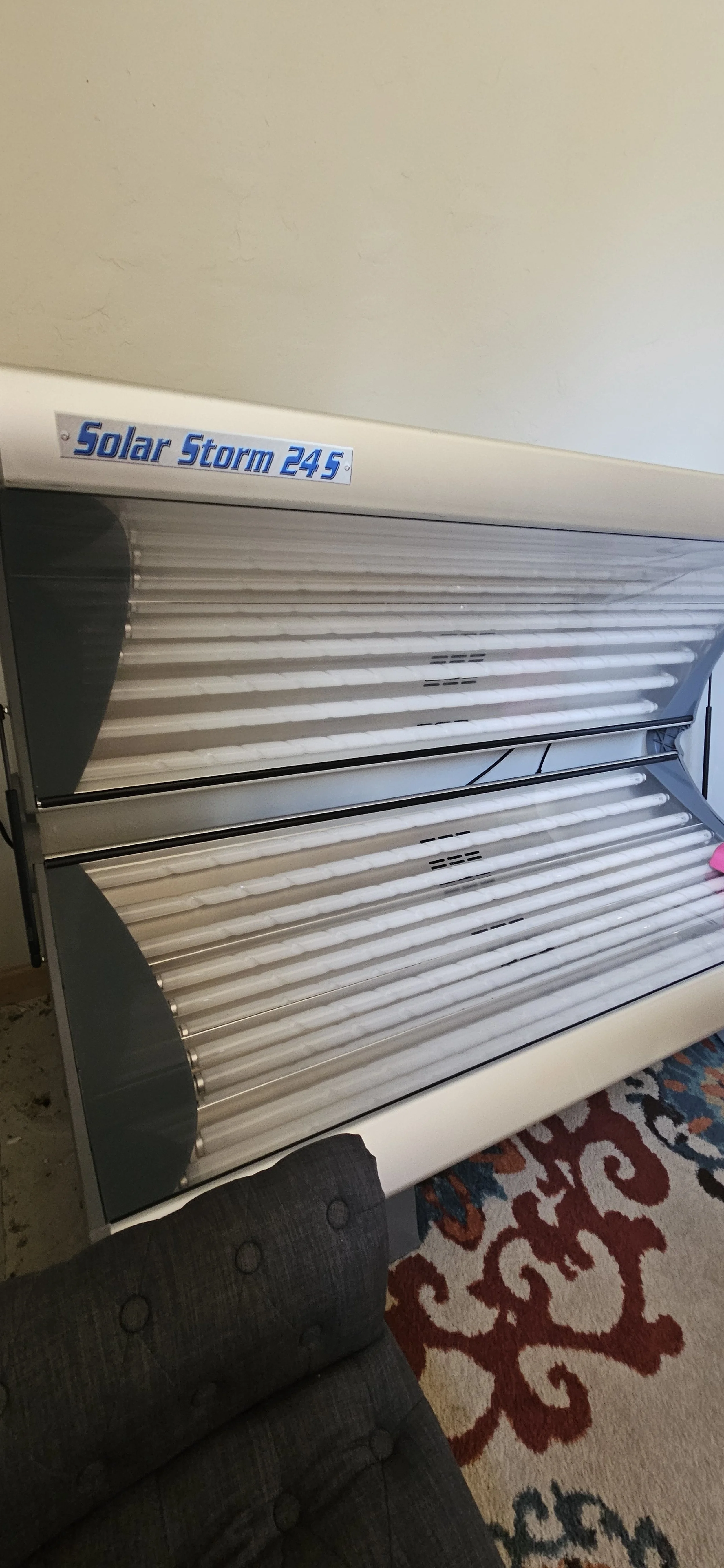 A clear Solar Storm 245 tanning bed with closed acrylics in a room with a patterned rug and a dark upholstered chair in the foreground.