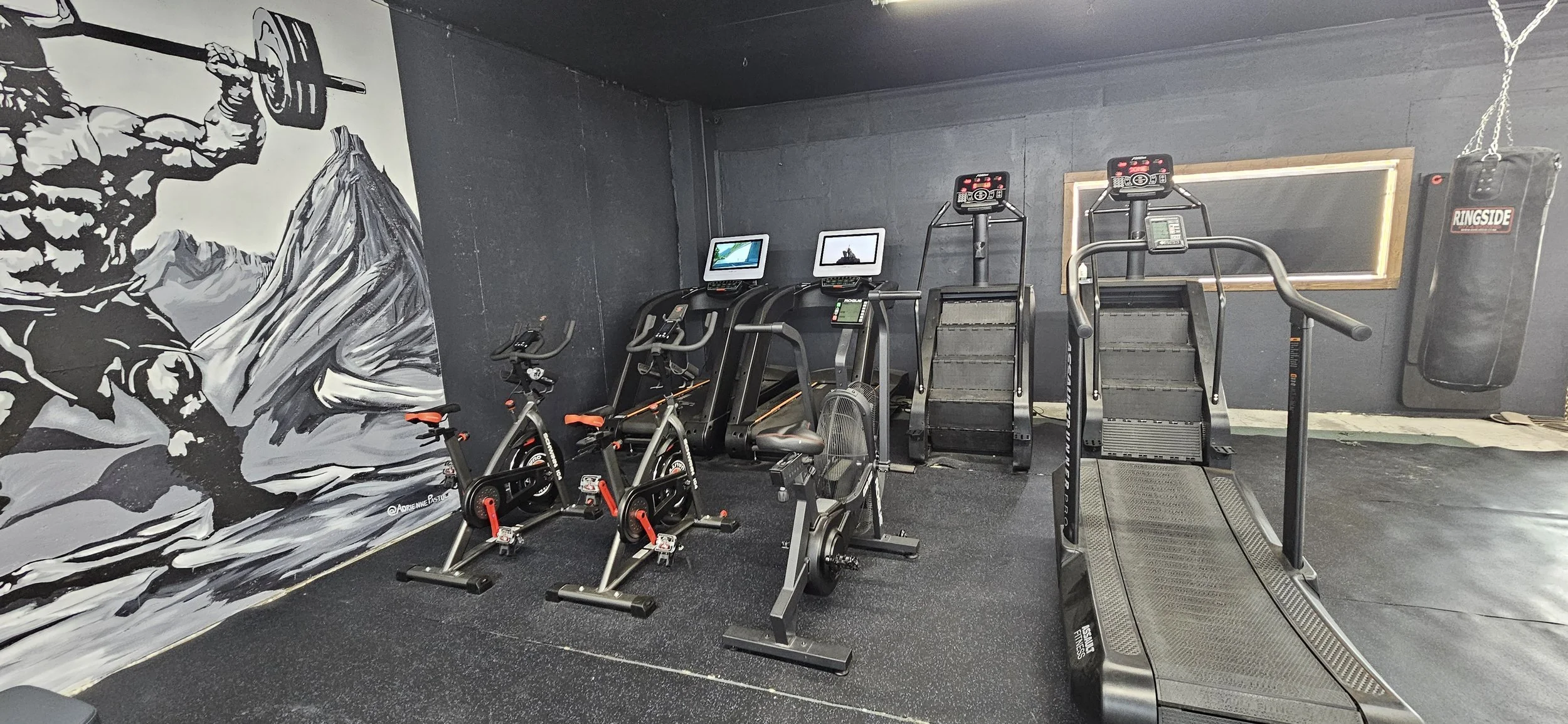 A gym with cardio machines, including two stationary bikes, two treadmills, and a stair stepper, with a punching bag hanging on the right side and a mural of an athlete lifting weights on the left wall.