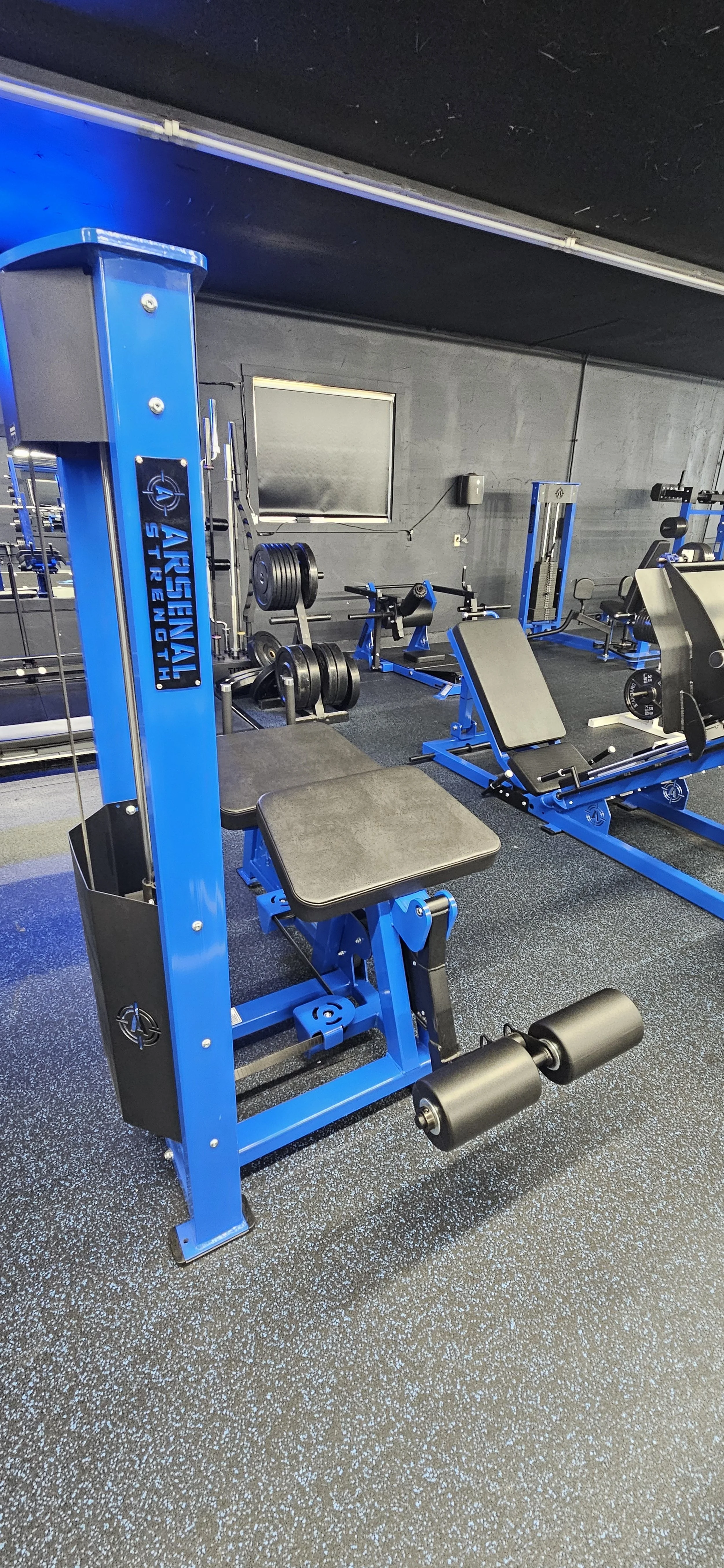 Blue and black exercise machine labeled 'Arsenal Strength' in a gym with various weights and workout equipment.
