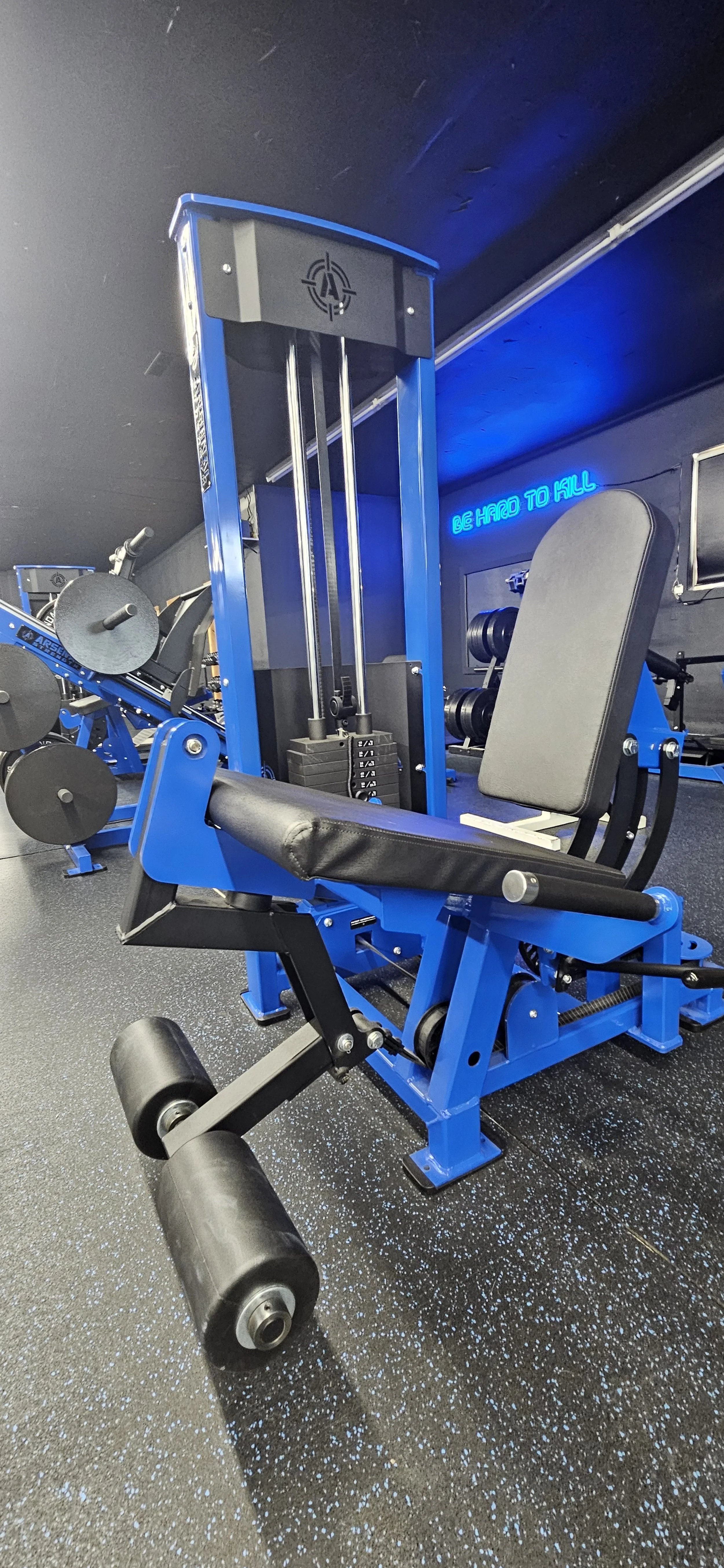 A gym with intensity training equipment, including a seated leg press machine and a weight sled with plates, set on a black and blue speckled floor. Blue neon sign in the background reads, 'BE HARD TO KILL.'