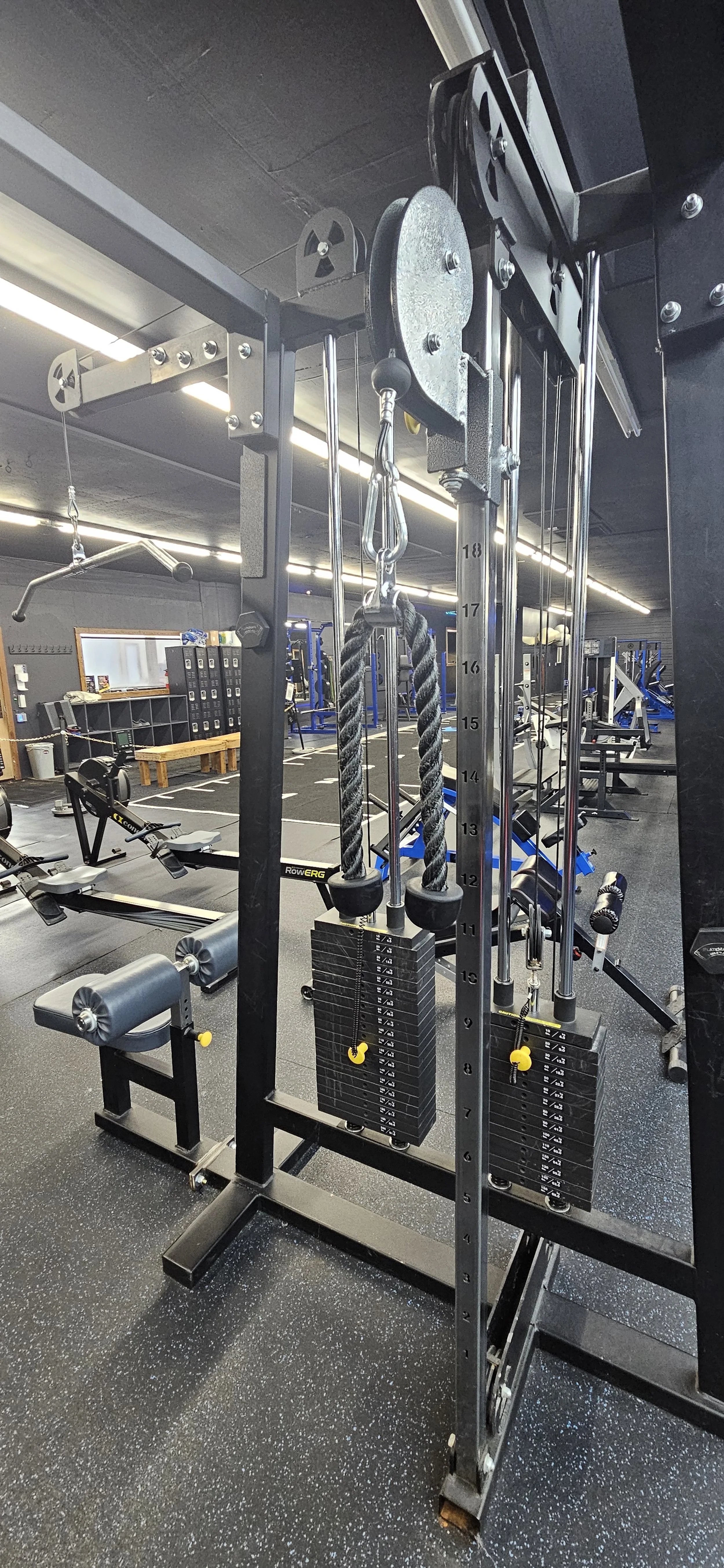 Gym equipment including a weight stack with ropes, rowing machines, and other workout stations in a fitness center.