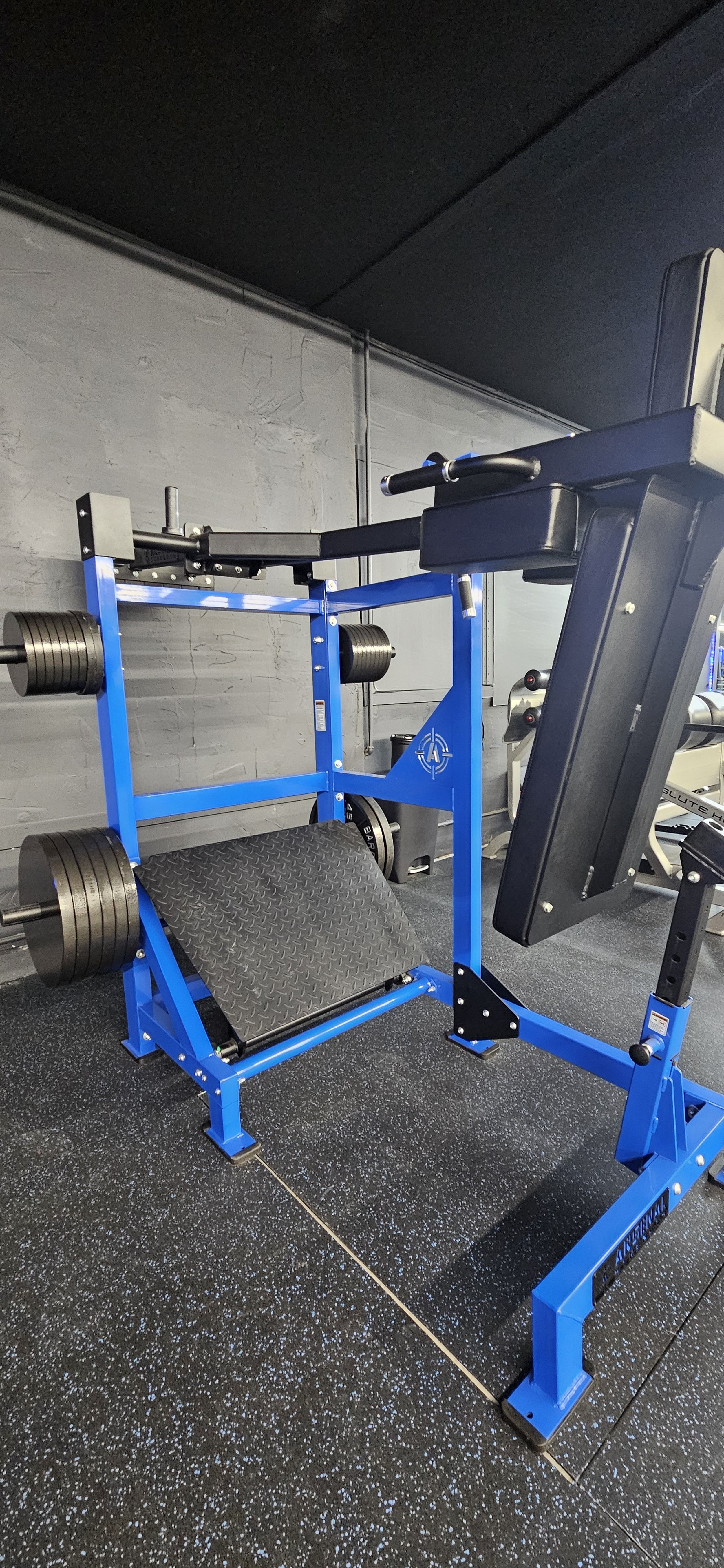 A blue and black weightlifting machine with weight plates on the sides, situated on a black rubber gym floor, against a gray wall.