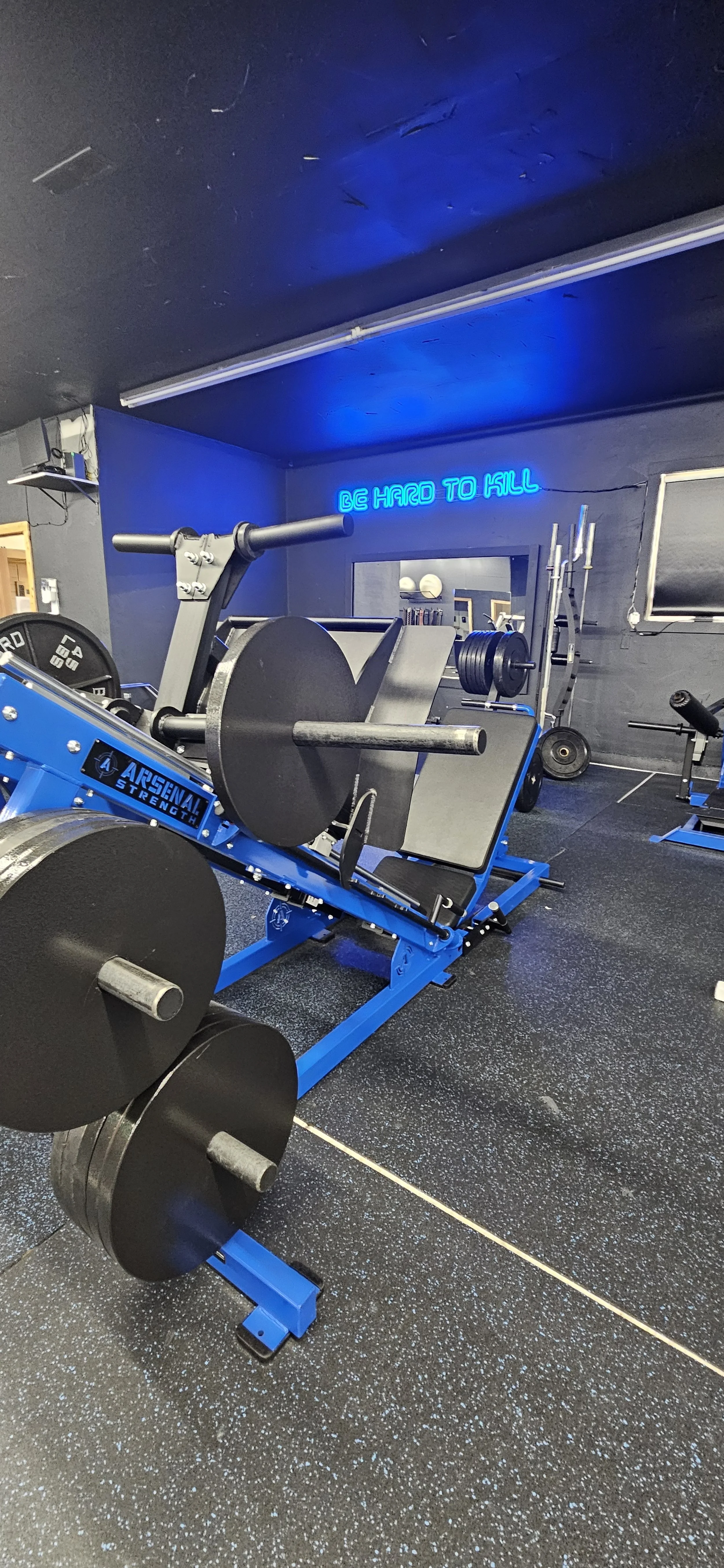 Gym with weightlifting equipment, including a blue ARSENAL strength machine, with black weights and barbells in the background. Neon sign on the wall reads 'BE HARD TO KILL'.