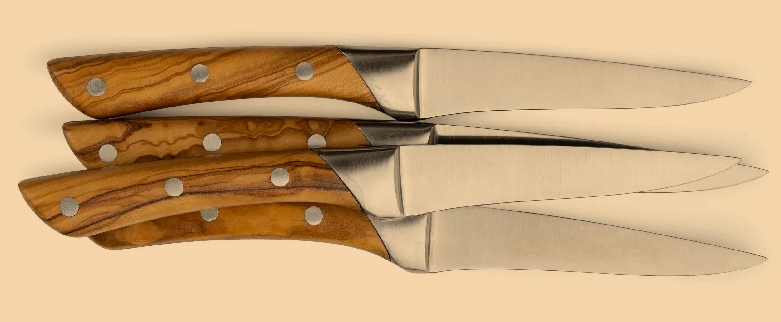 Three sharpened kitchen knives with wooden handles and stainless steel blades, arranged horizontally on a beige background.