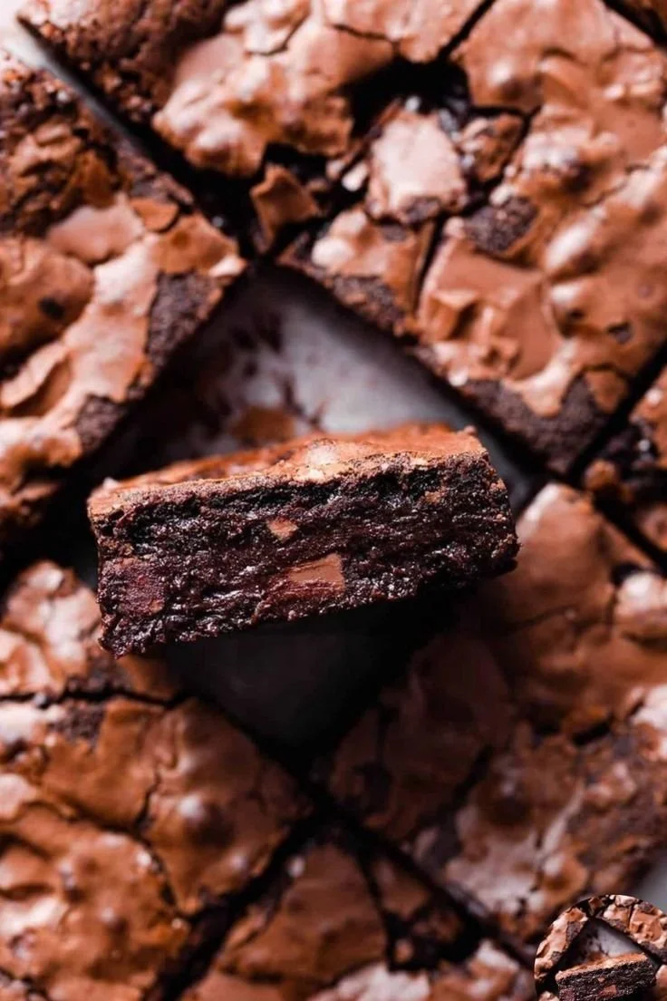 Close-up of a stack of brownies with a single piece prominently displayed in the center, showing a moist, fudgy interior and a cracked, slightly crispy top.