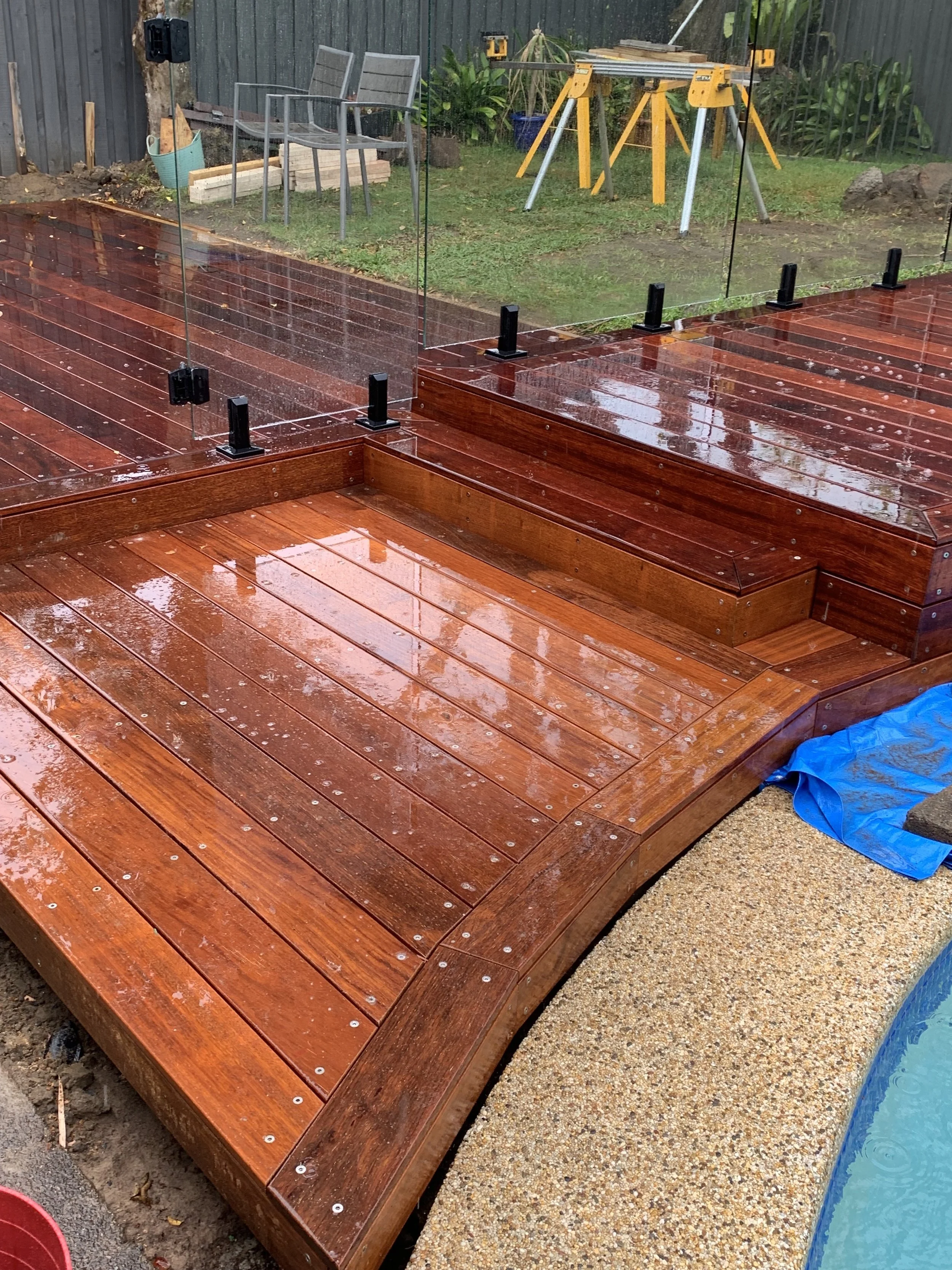 Rain-soaked wooden deck with glass panels and black brackets, adjacent to a concrete pool edge, in a backyard with gardening tools and outdoor furniture.