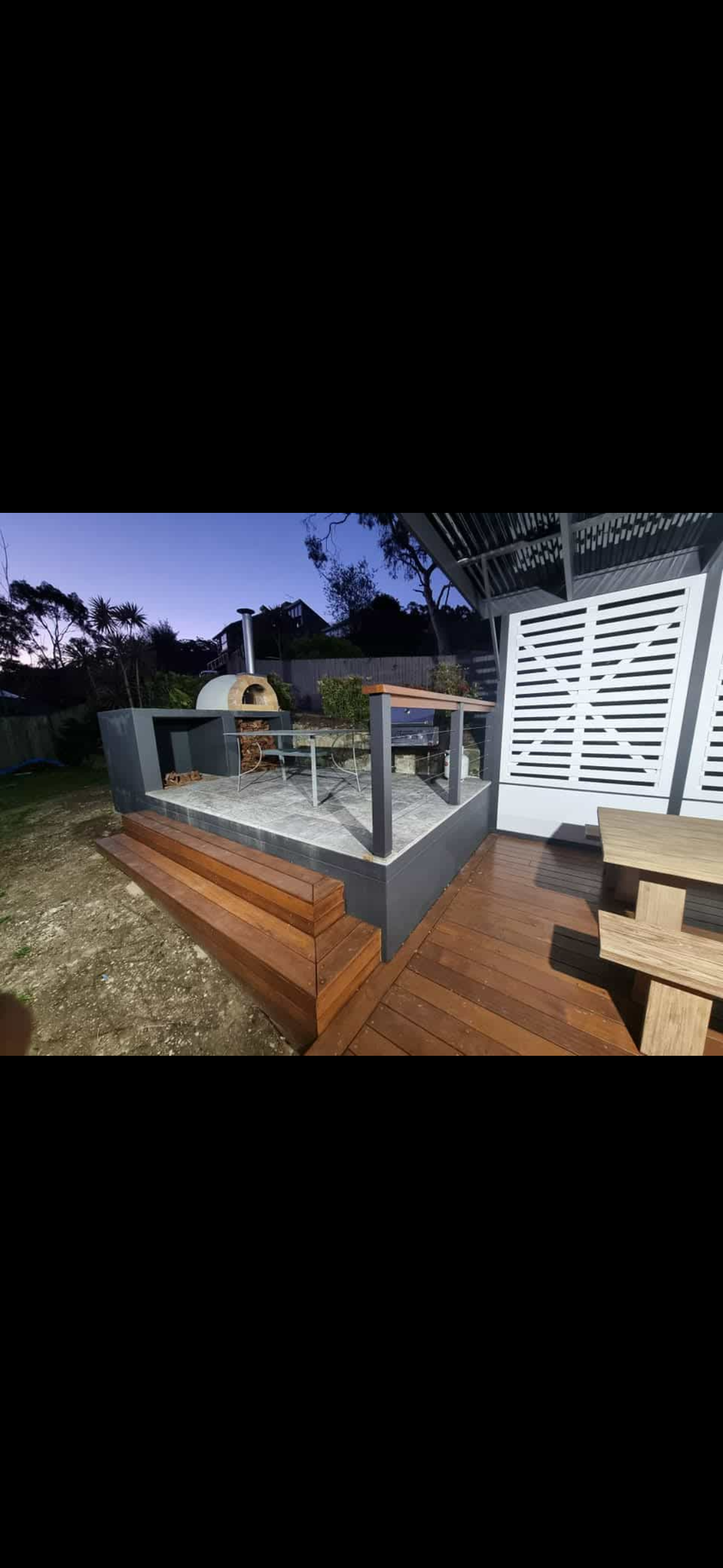 Outdoor patio area with wood and tiled flooring, a built-in pizza oven, a grill, and outdoor furniture, surrounded by a fence and trees, during dusk.
