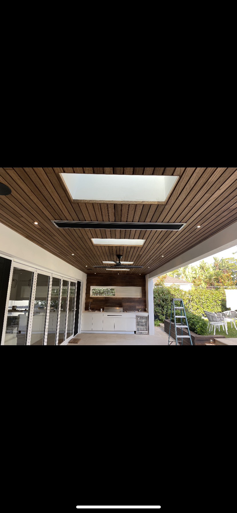 Outdoor patio with wooden ceiling, skylights, and ceiling fan, featuring sliding glass doors, a built-in outdoor kitchen, and garden area with trees and outdoor furniture.