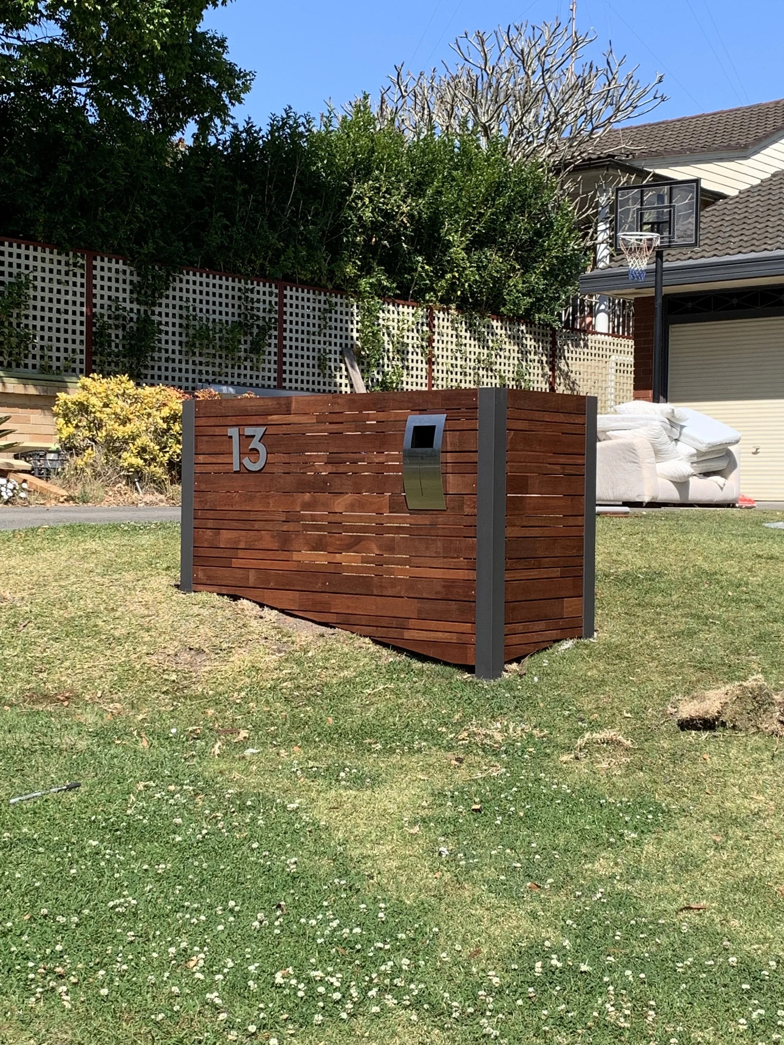 A modern wooden fence with house number 13, a mailbox, and a mailbox slot. In the background, a yard with a basketball hoop, a couch, and some backyard plants and trees.