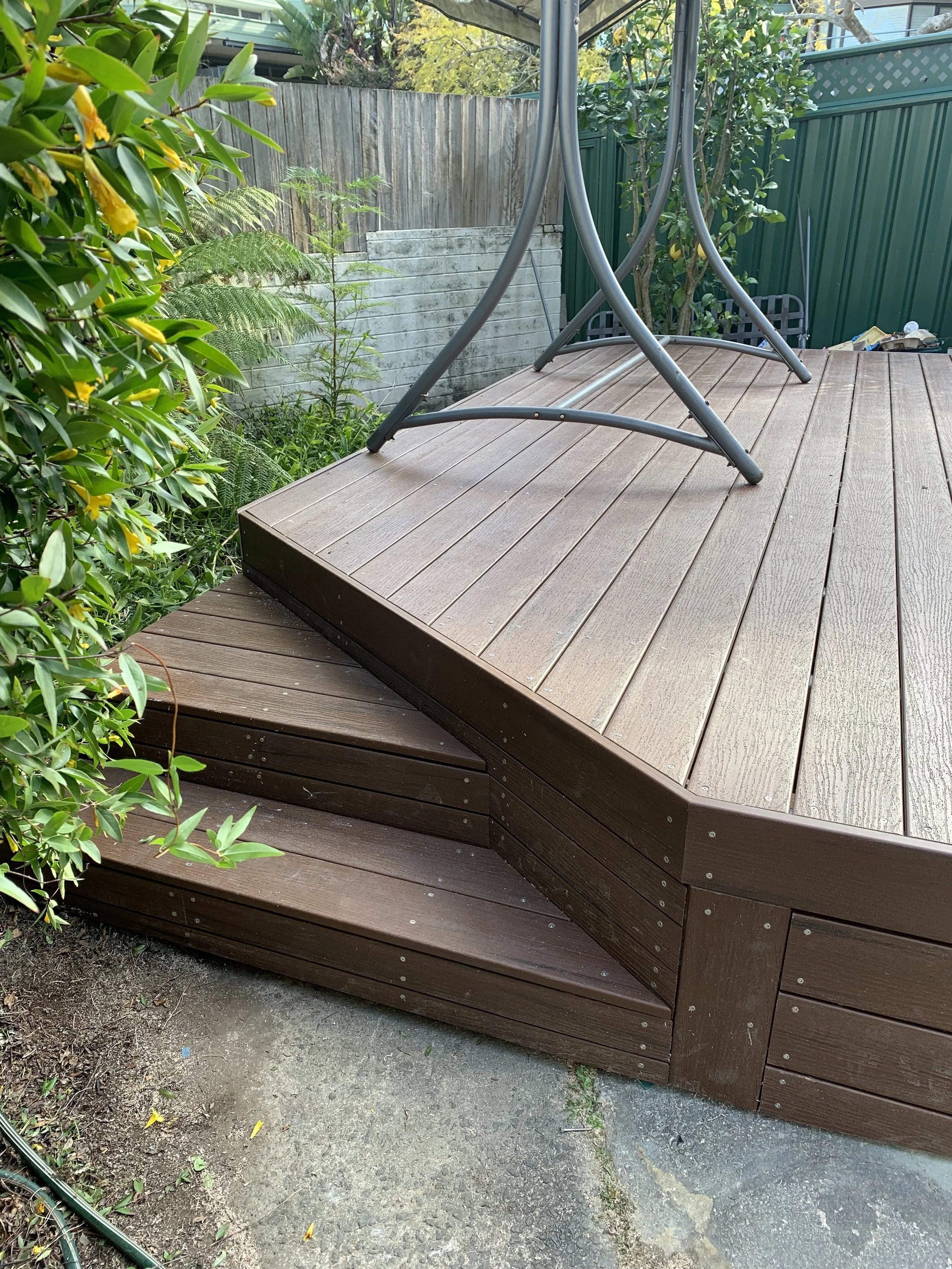 A wooden outdoor deck with a metal table upside down on top of it, adjacent to green plants and a garden fence.