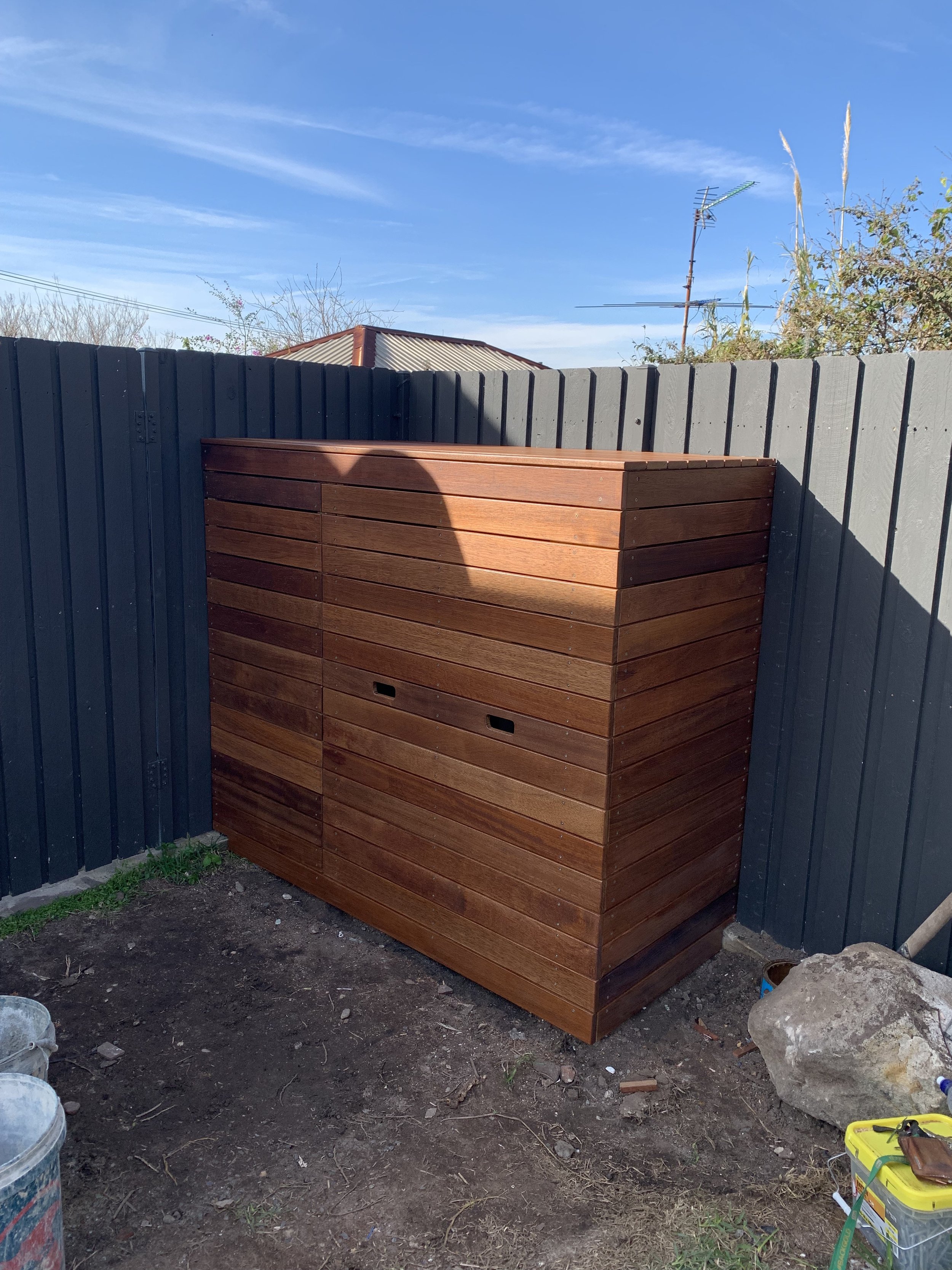 A wooden storage shed with horizontal slats, situated against a dark gray fence, under a blue sky with some clouds and a TV antenna visible in the background.