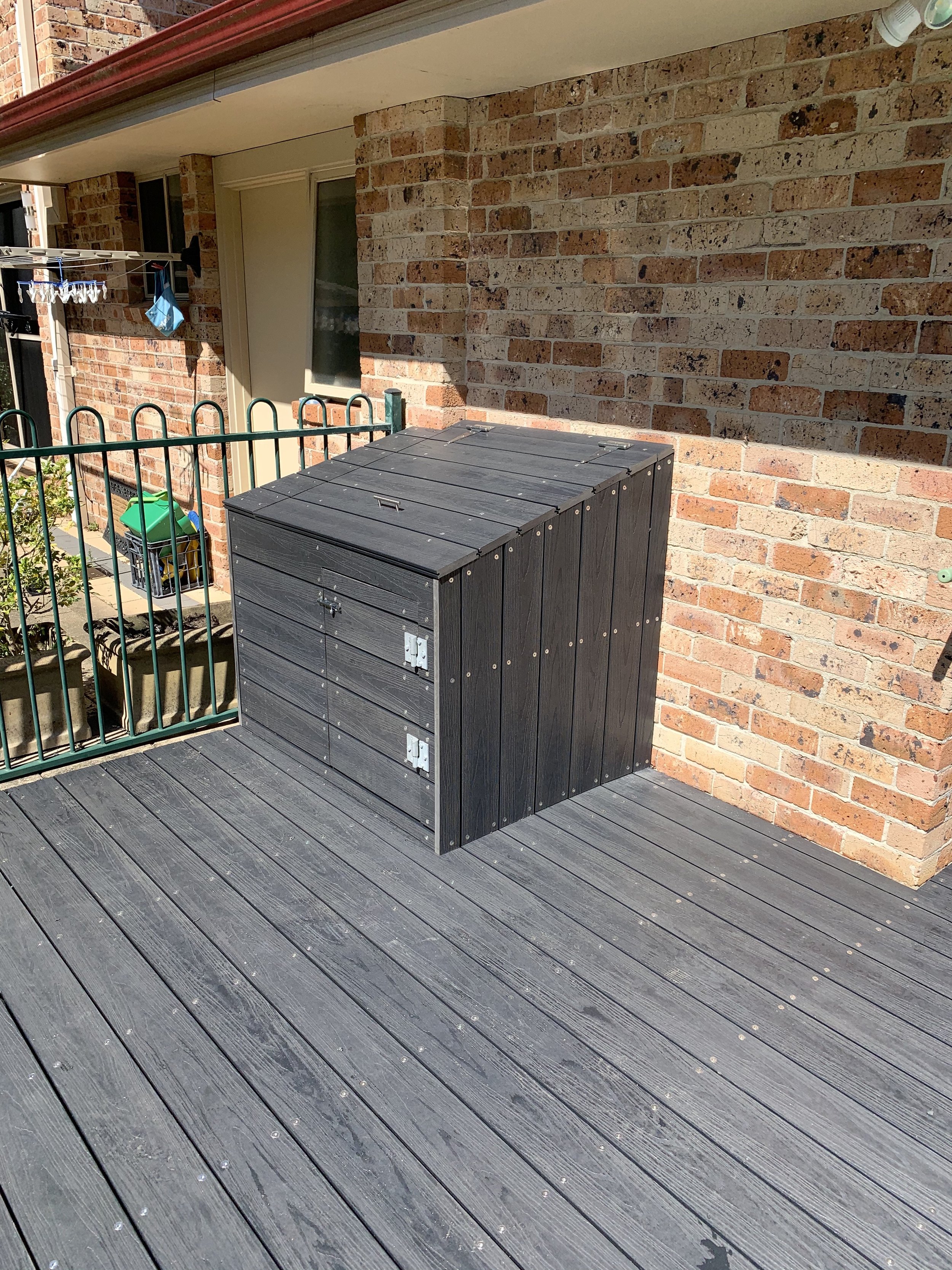 A small outdoor deck area with wooden flooring and a brick wall. There is a black storage box with a lid, positioned near the wall. A green fence surrounds part of the deck. A drying rack with some clothespins, a blue cloth, and a small box are visible in the background.