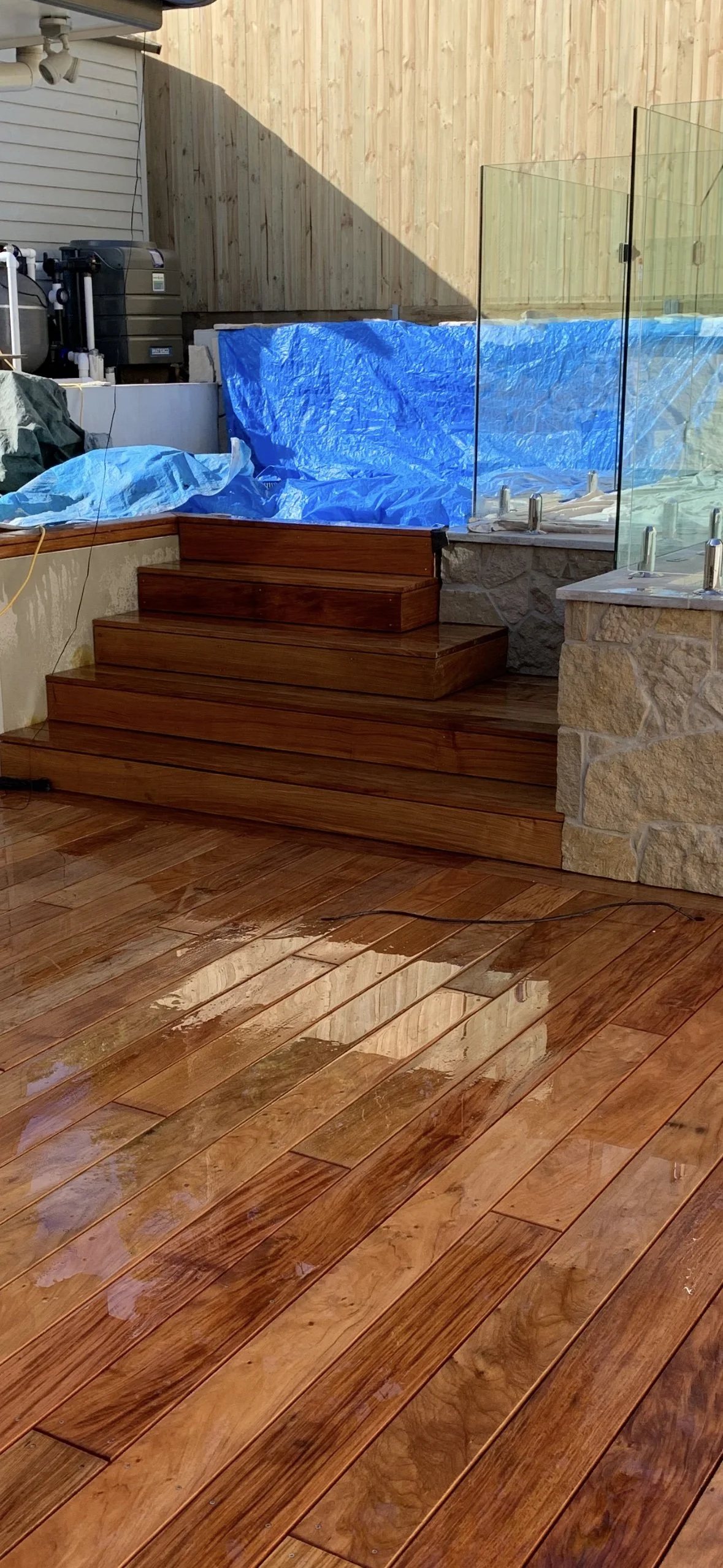 Wooden stairs and deck area, with a partially covered hot tub surrounded by a stone wall and glass fencing, and construction materials in the background.