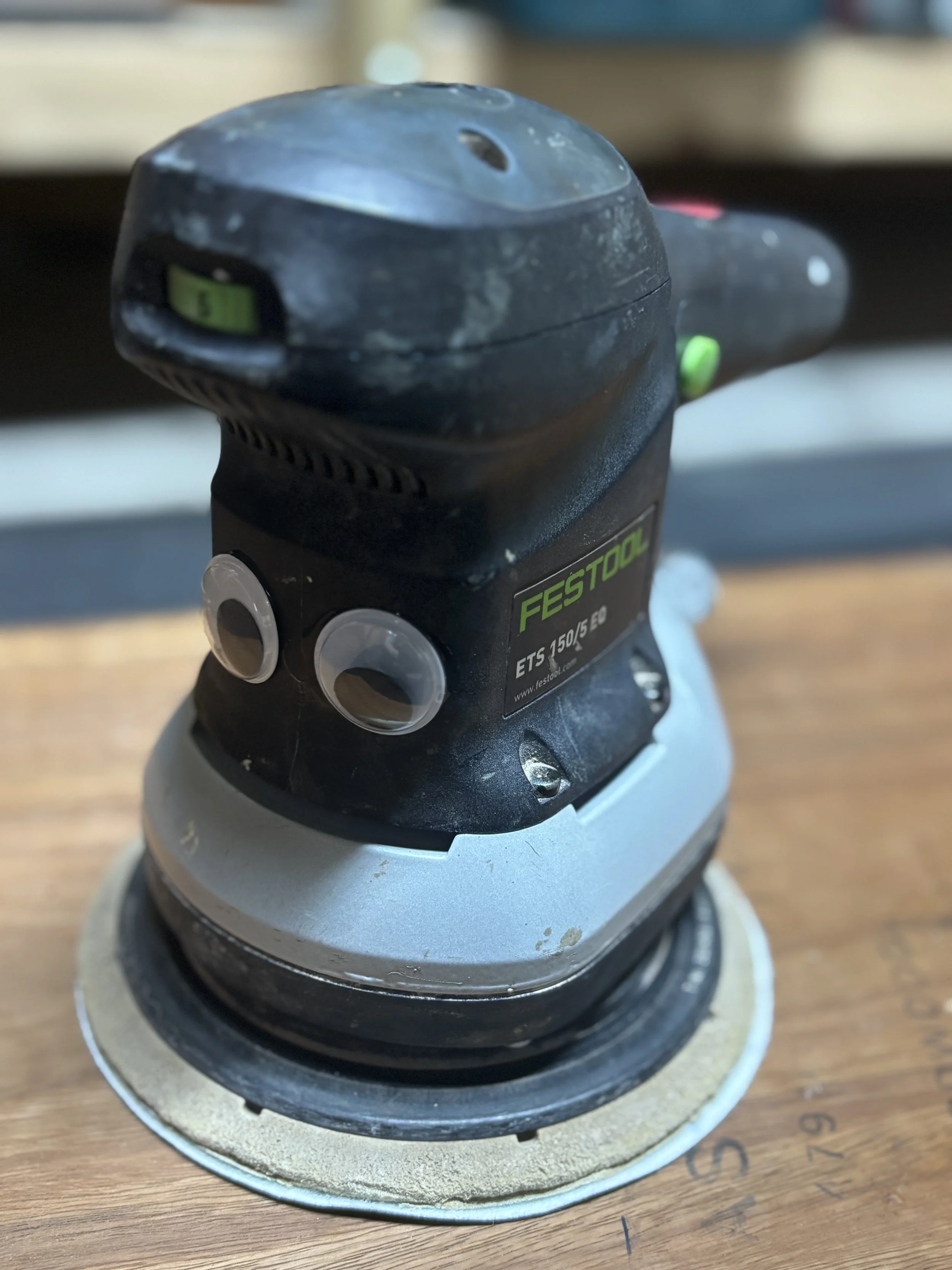 Close-up of a black Festool orbital sander with a corded attachment, resting on a wooden surface.