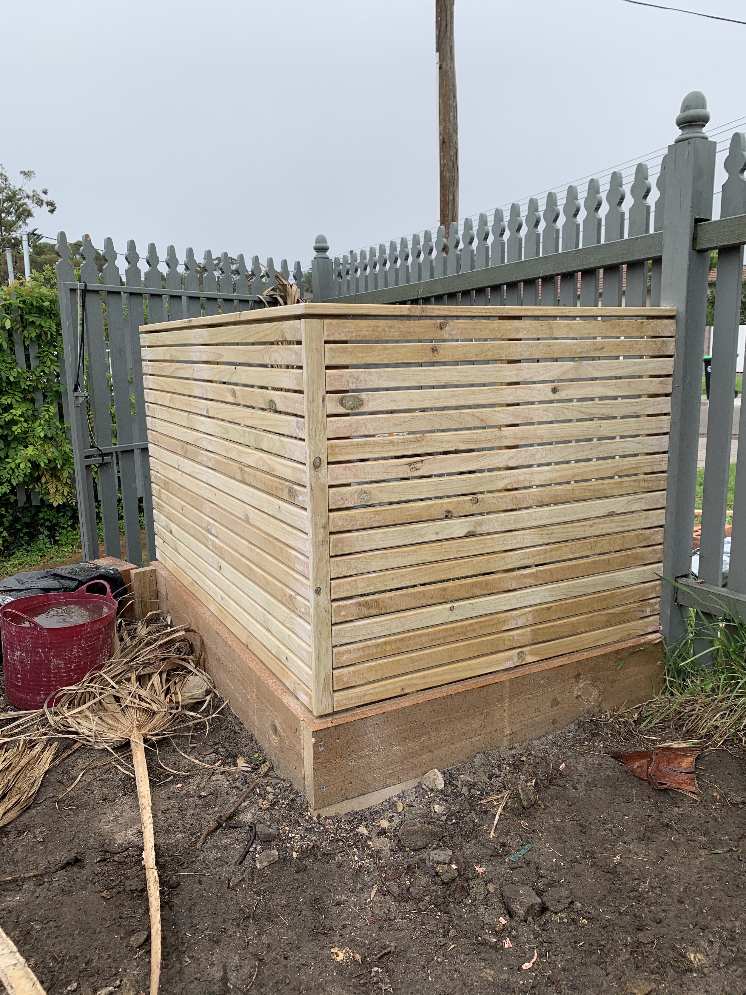 A partially built wooden privacy screen or fence attached to a concrete foundation near a gray fence, with a red bucket and construction tools nearby, on a dirt ground outdoors.