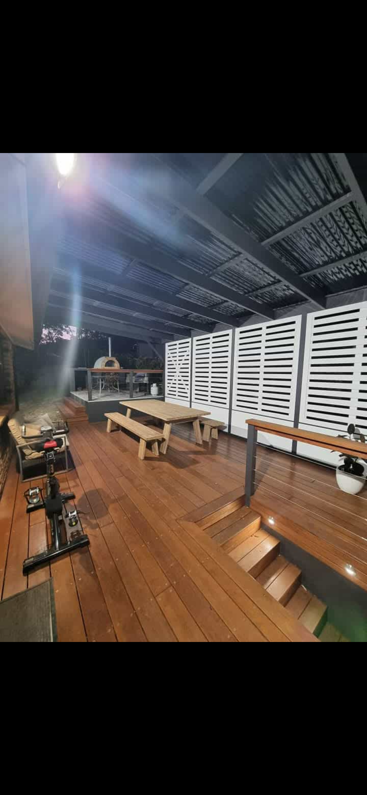 Outdoor patio with wooden decking, picnic table, a grill, stairs leading down to a swimming pool, and a white privacy screen. Nighttime setting.