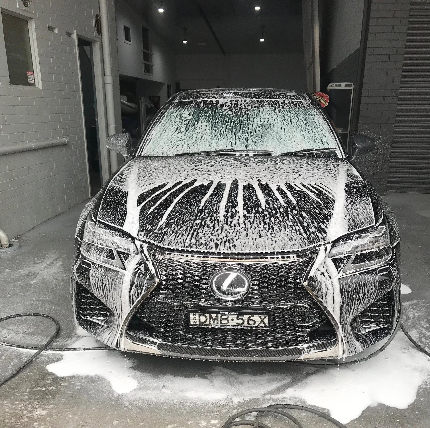 Black Lexus car being washed with soap and foam inside a garage. The front of the car is covered in white soap suds, with a lather pattern on the hood and windshield. Autoprep Sydney Car Detailing -