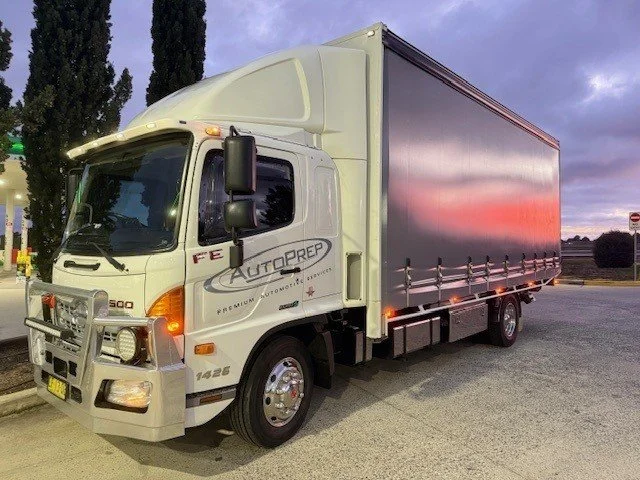 A white medium-sized delivery truck parked on a paved area, under a slightly cloudy sky, with trees in the background.