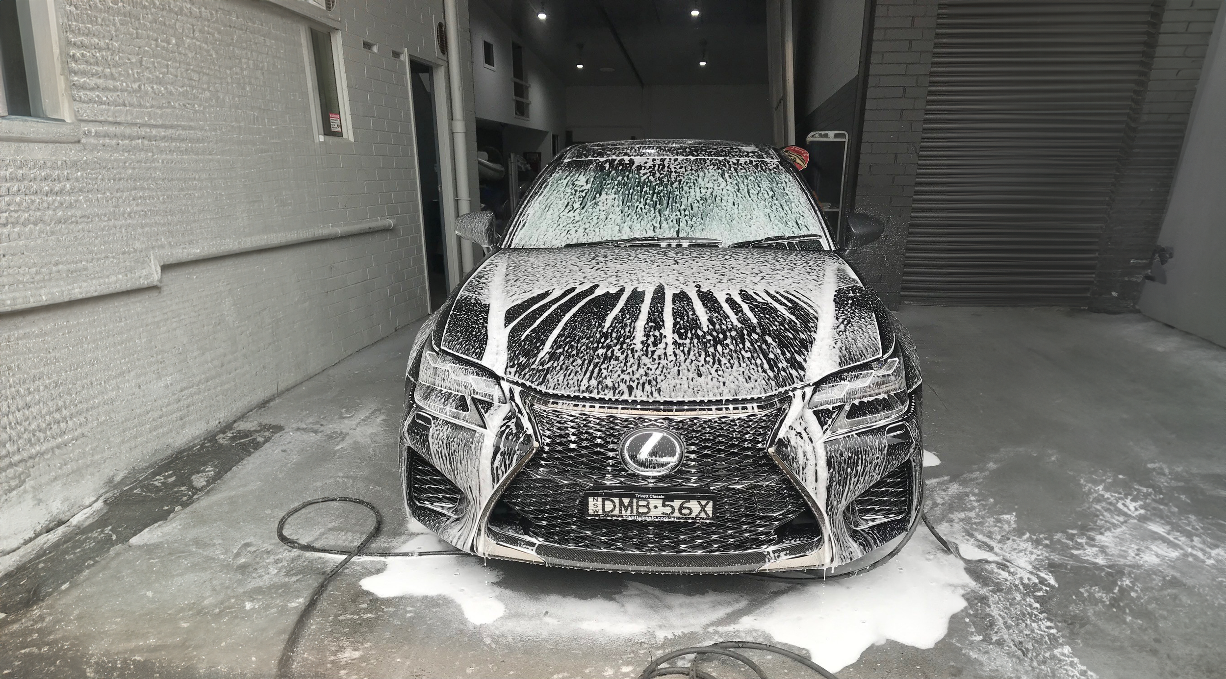 Autoprep Sydney Car Detailing - Black Lexus sedan being washed in a car wash bay, covered in white soap suds.