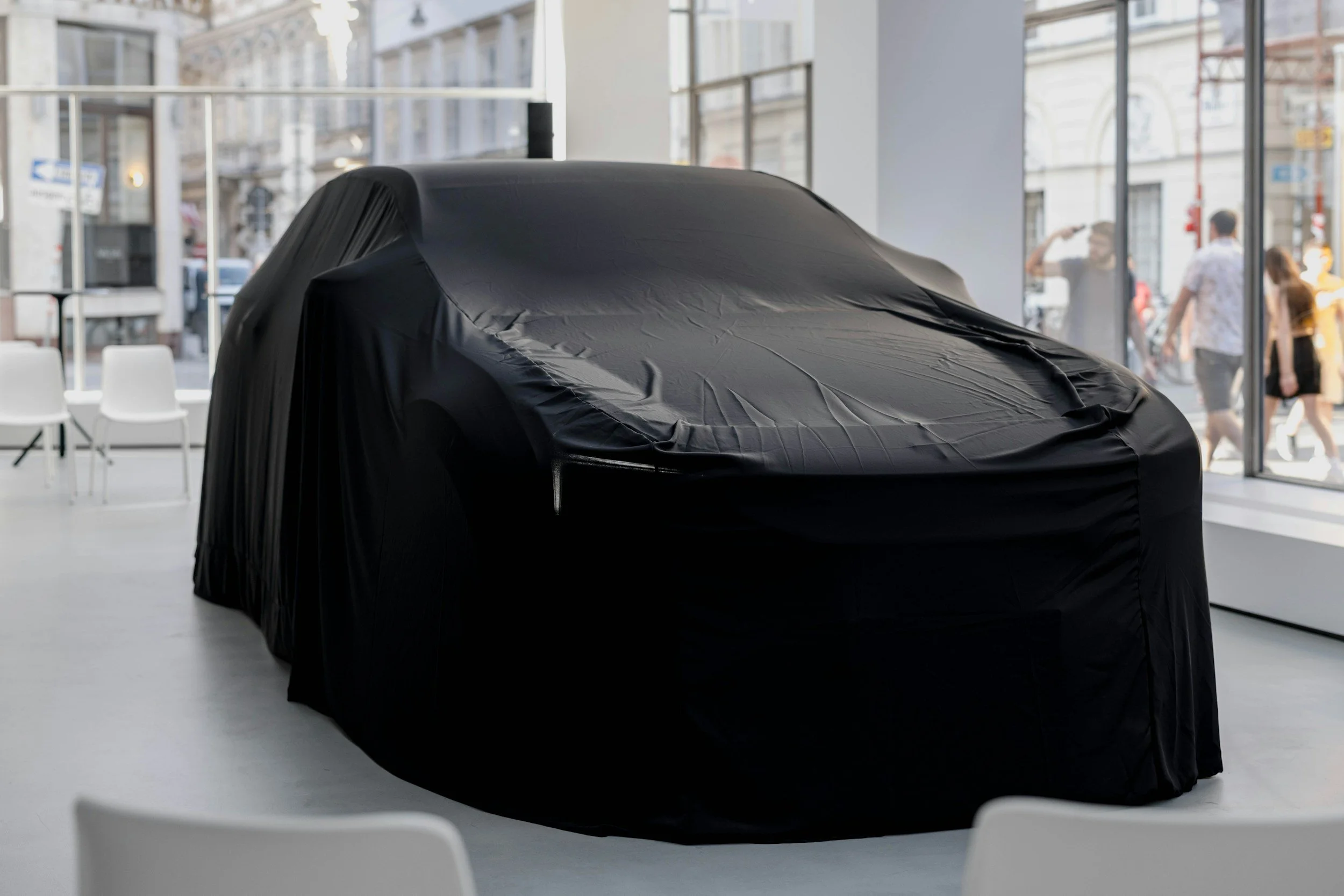 A car covered with a black cloth, inside a modern showroom with large windows and white chairs.