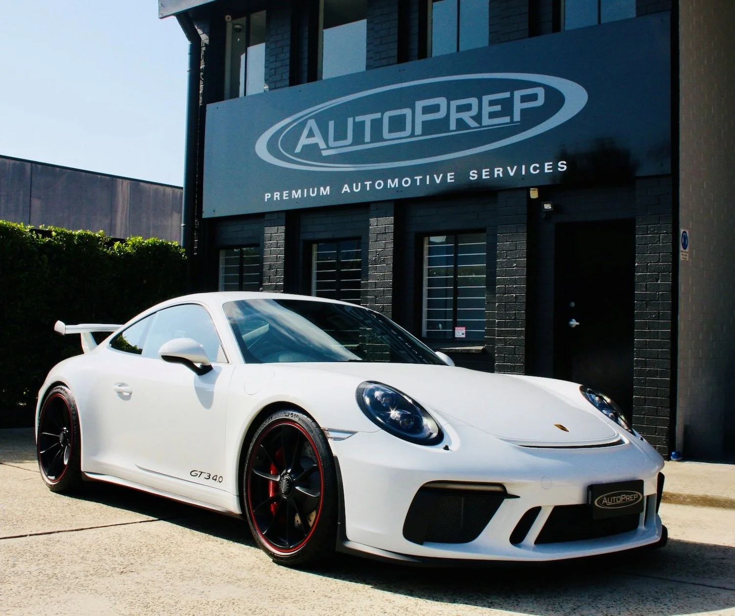 A white Porsche 911 GT3 with red accents parked outside an automotive service shop named AutoPrep.