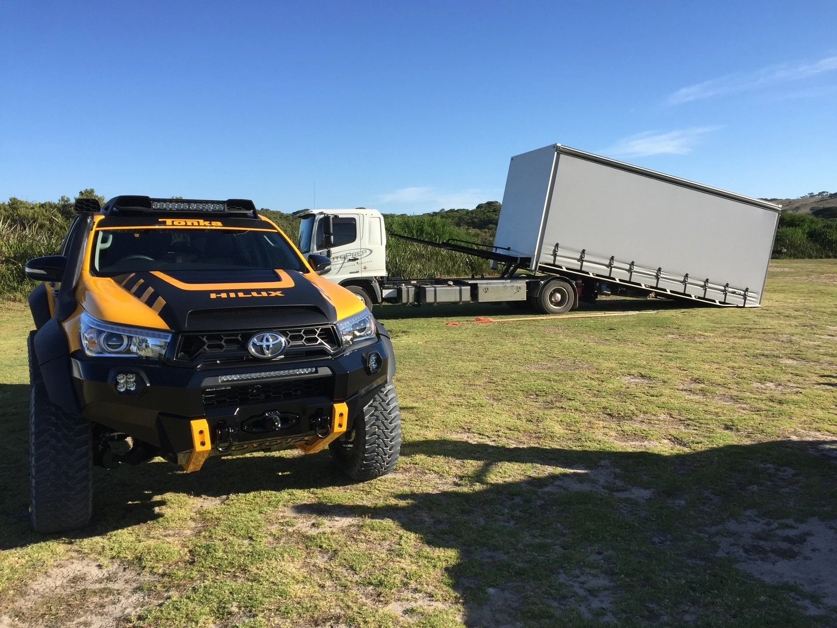 A yellow and black Toyota Hilux with off-road modifications parked on grassy terrain, next to a white flatbed truck with a tilted cargo area.