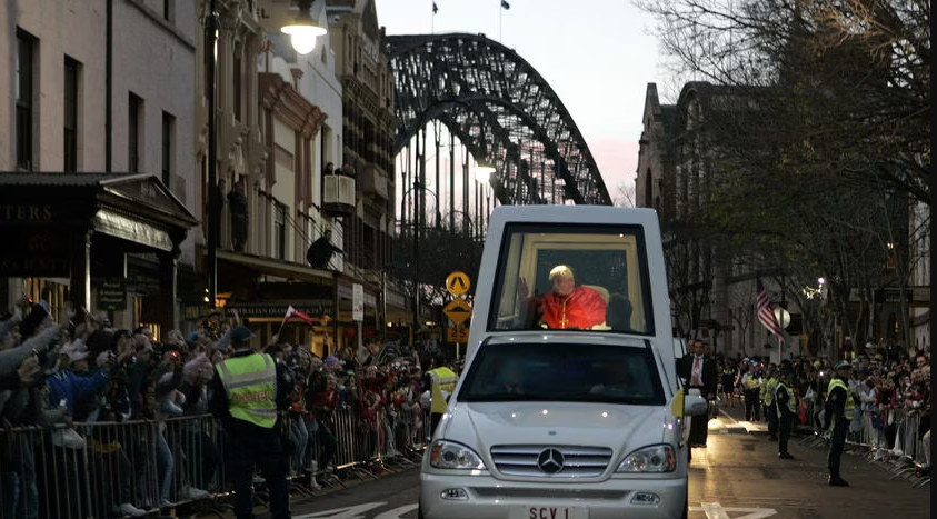 A parade procession on a city street featuring a person dressed as Santa Claus riding in an open vehicle. The street is lined with spectators behind barriers and security personnel. There is a large arch structure in the background and American flags are visible.