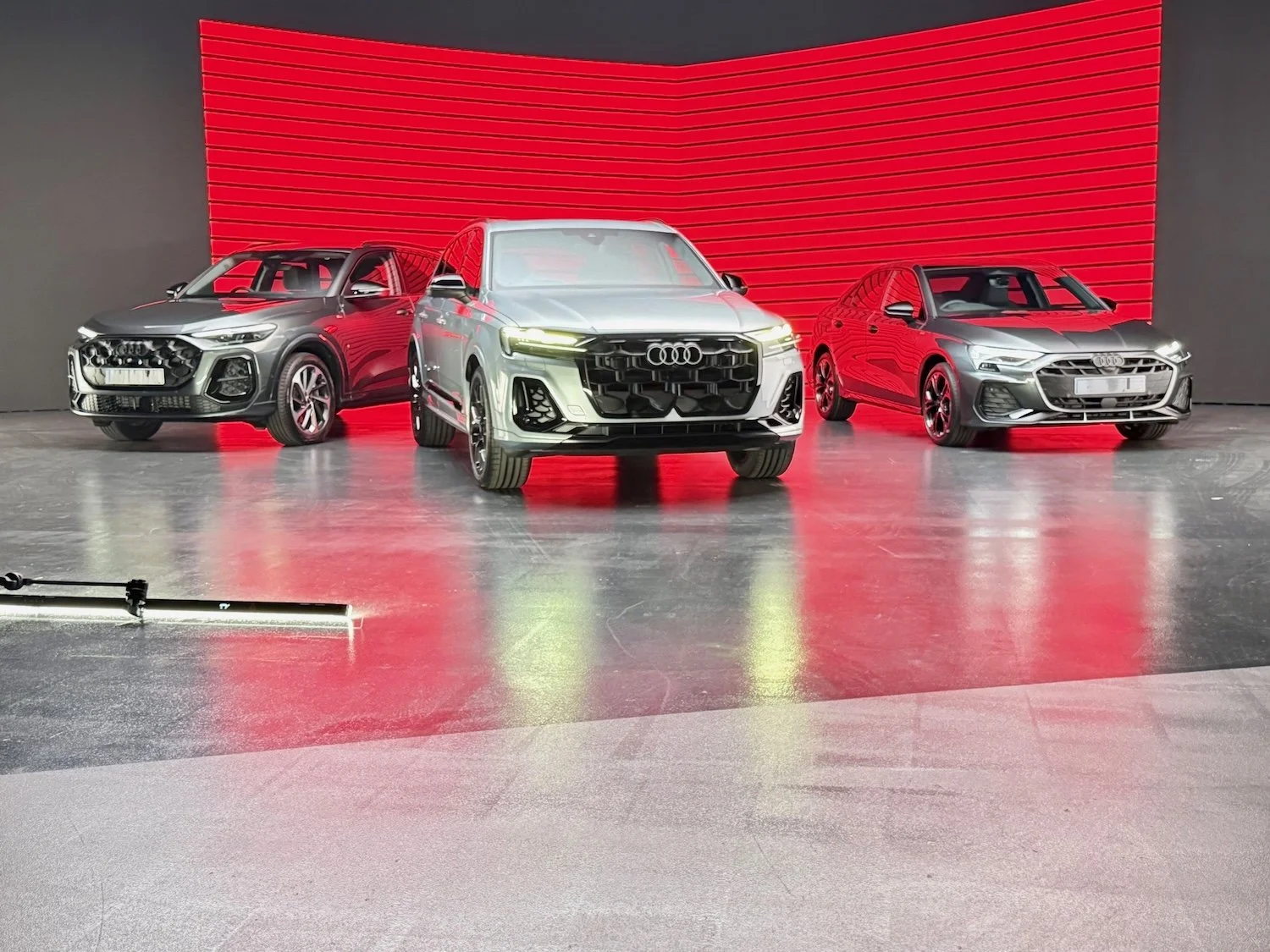 Three new Audi cars are displayed in front of a red and black background at an auto show or car unveiling event.