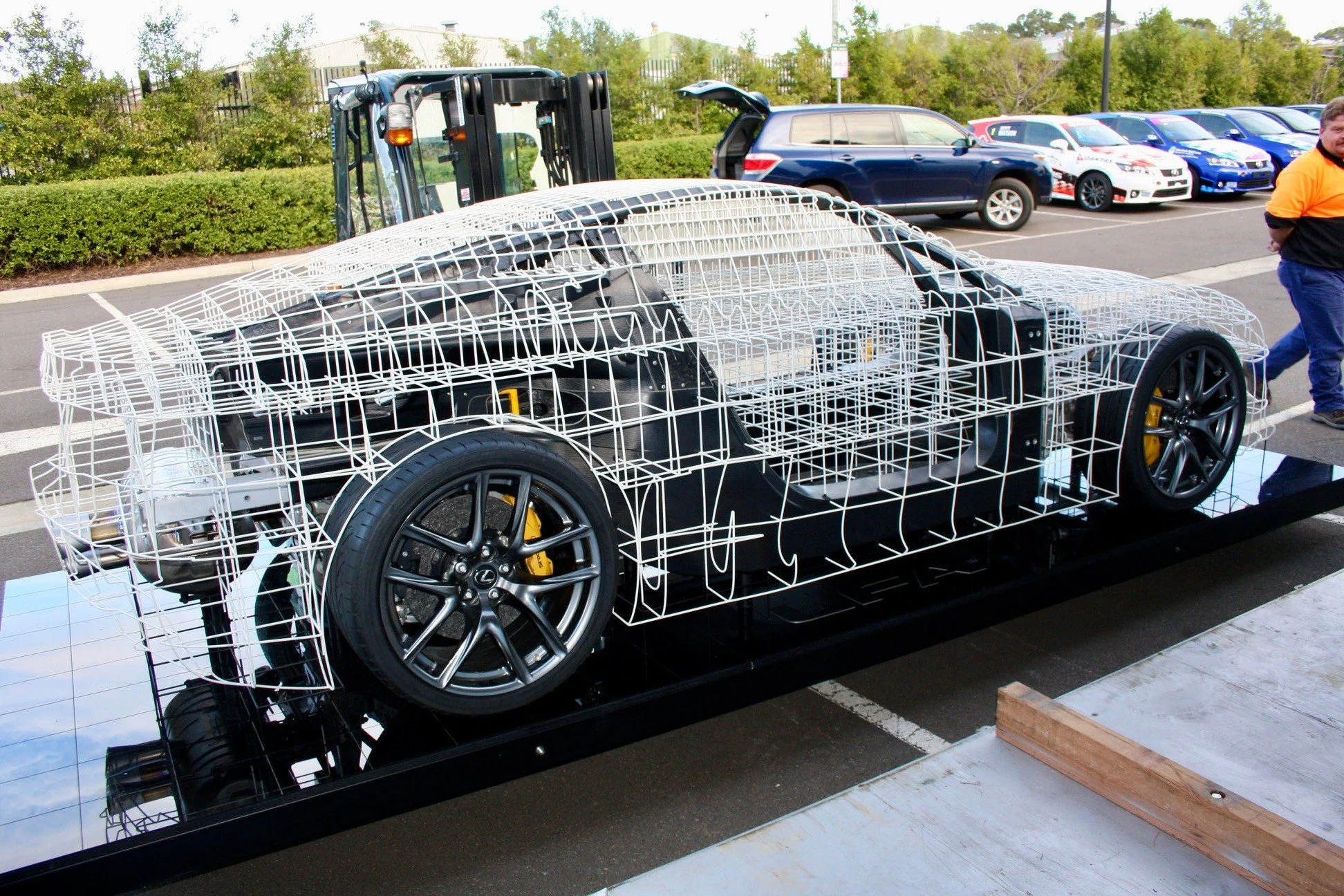 Prototype car frame constructed with white wireframe structure, placed on a black platform in an outdoor parking lot, with parked cars and trees in the background.