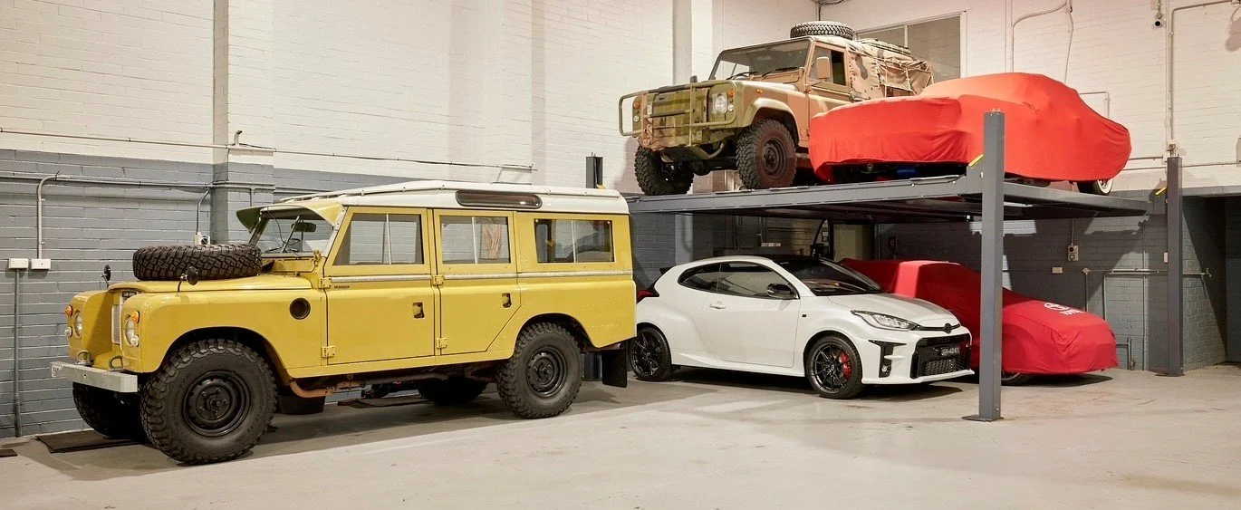 An indoor garage with two cars on the ground level and three cars on the upper level, including a yellow vintage off-road vehicle, a white sports car, and three other vehicles covered with red car covers.
