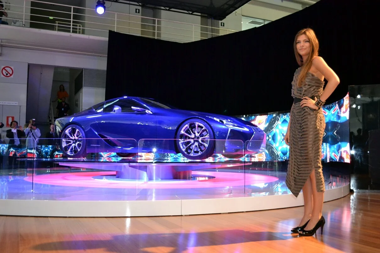 A woman in a patterned dress and high heels stands next to a sleek blue sports car on display at an auto show.