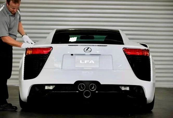 Rear view of a white Lexus LFA sports car with a person inspecting it, in front of a gray industrial-style garage door.