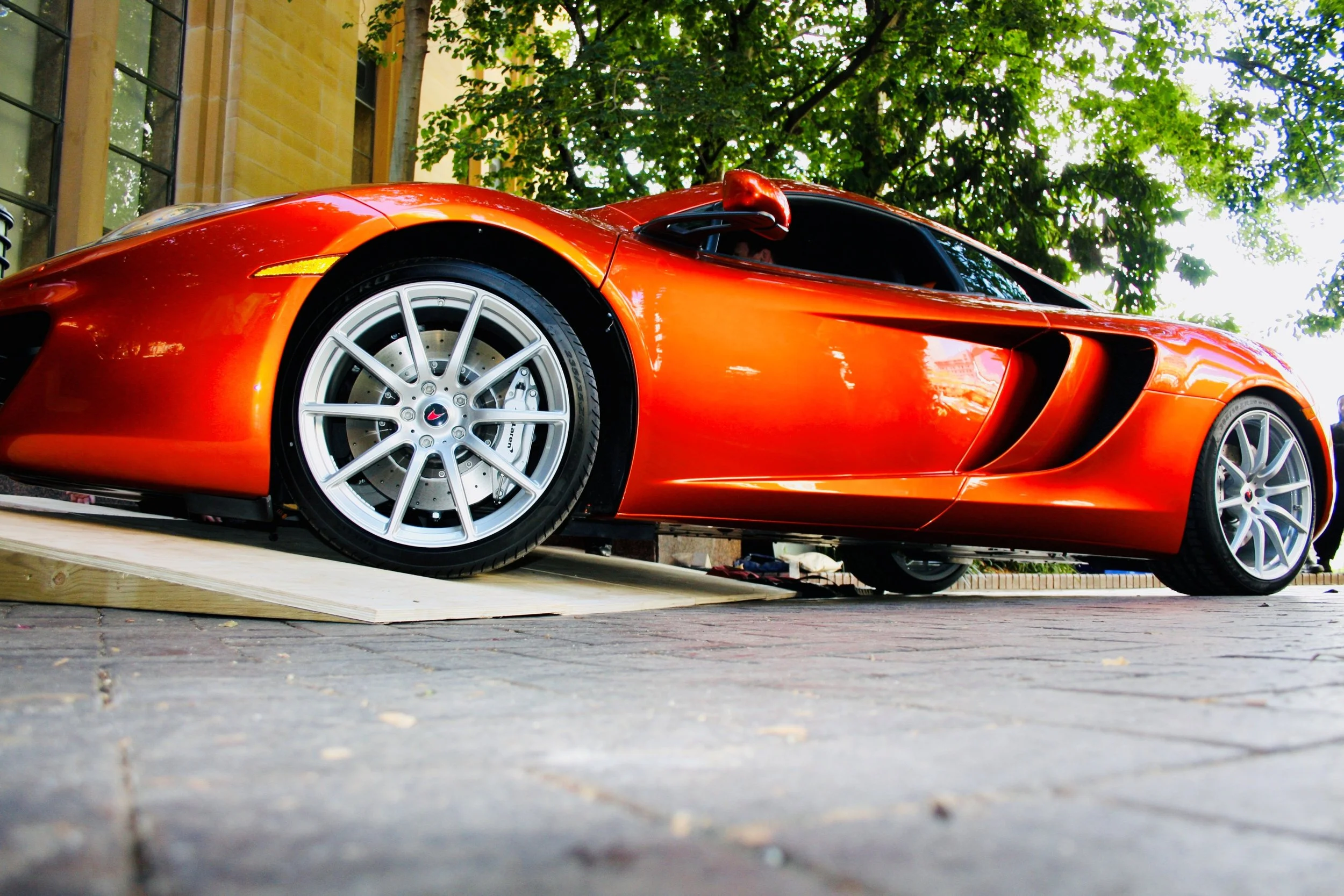 Low-angle view of an orange sports car parked on a brick pavement with trees and a building in the background.