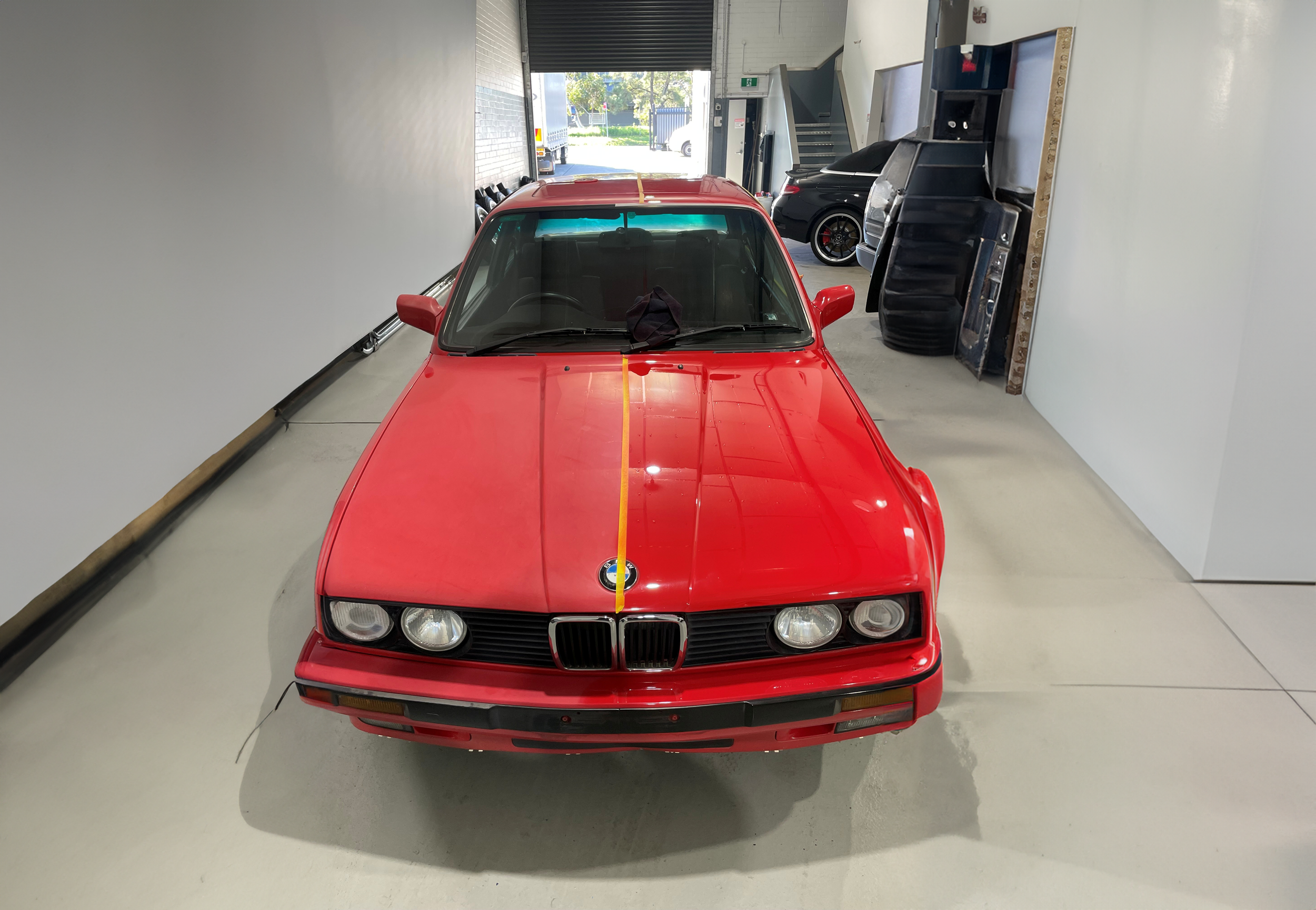 Front view of a red vintage BMW sports car parked indoors, with a black car and equipment in the background.