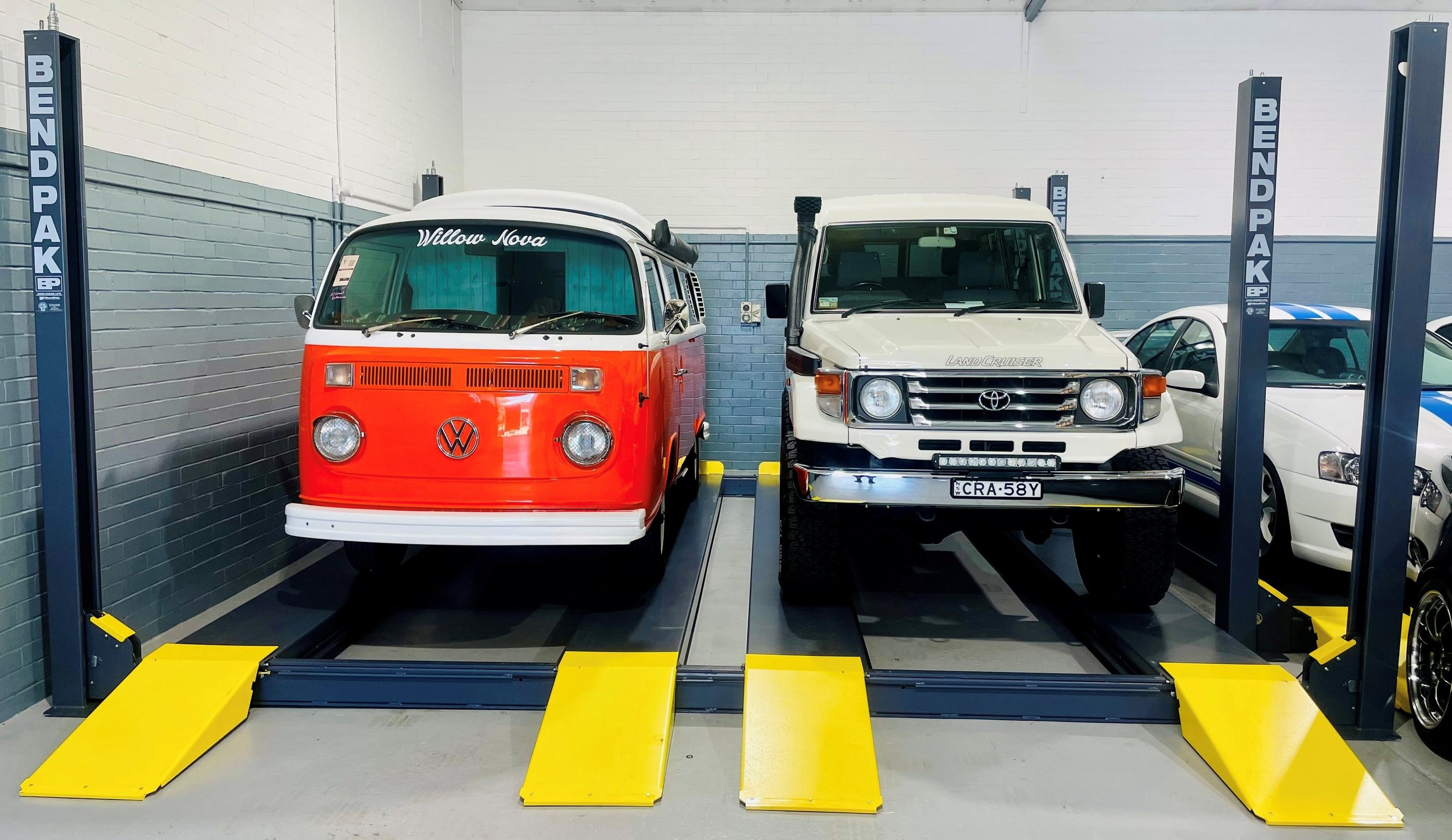 Two vintage cars, a Volkswagen van and a Toyota Land Cruiser, parked on vehicle lifts in a garage.