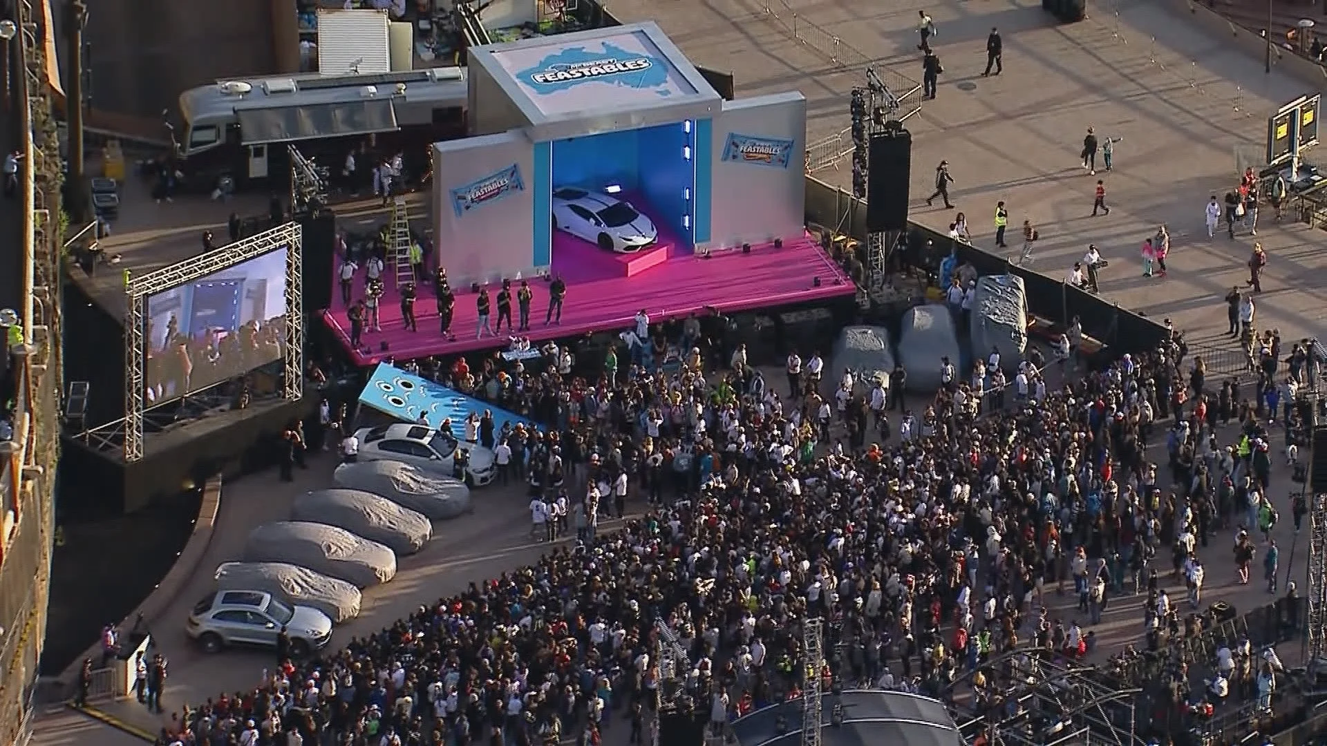 An outdoor event with a stage, a large crowd, and a white sports car displayed on the stage. The crowd is gathered in front of the stage, and there are several parked cars nearby.