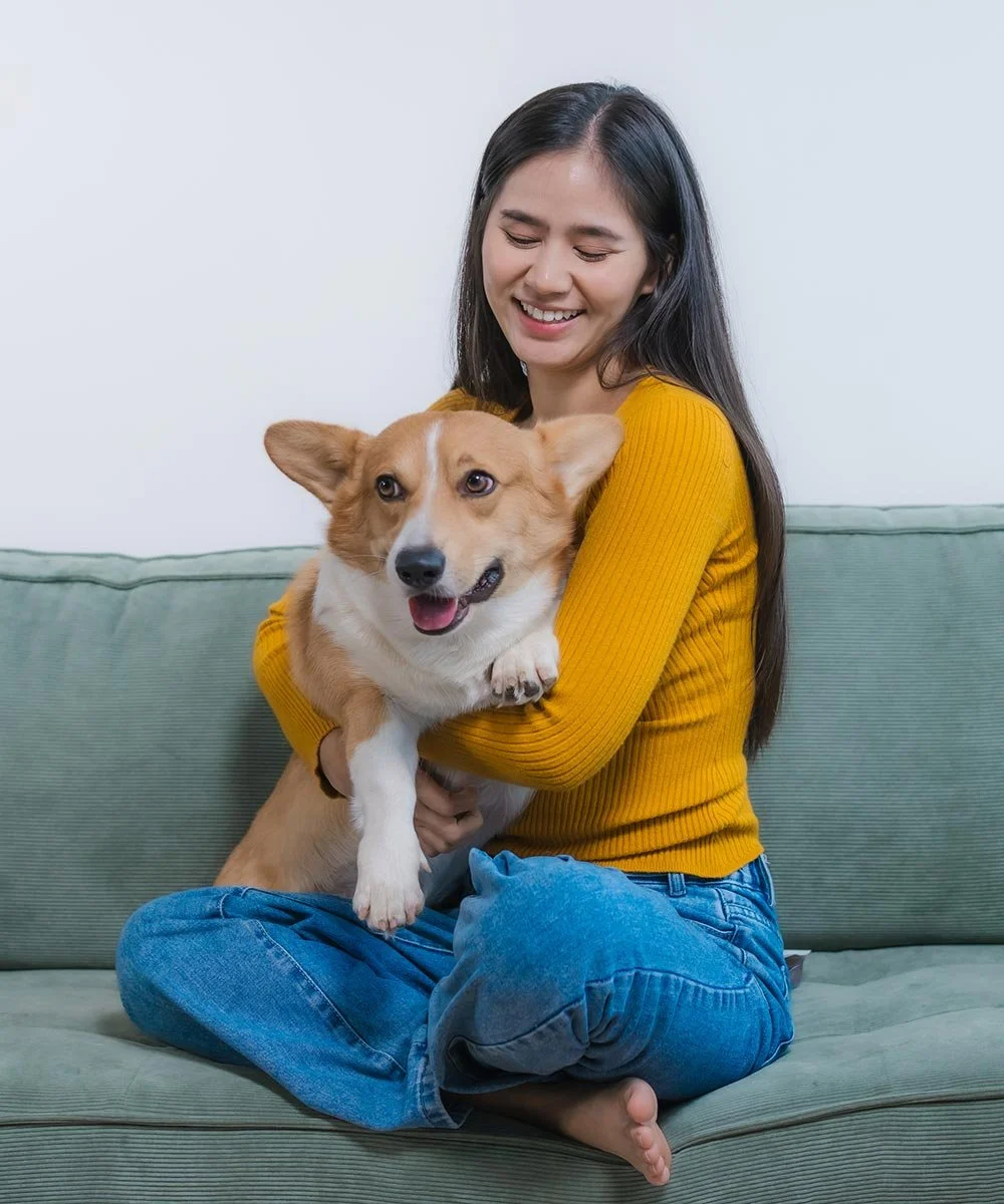A woman in a yellow sweater sitting cross-legged on a green couch, smiling and holding a joyful corgi dog.