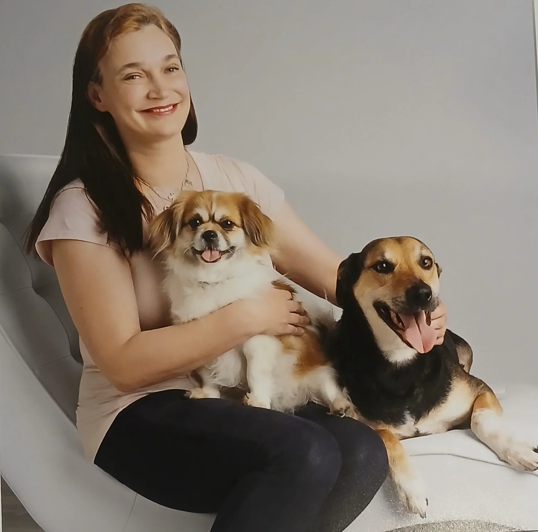 A smiling woman with long brown hair sitting on a white chair, holding two dogs, one small and fluffy and the other larger with a black and tan coat, both dogs appear happy and content.
