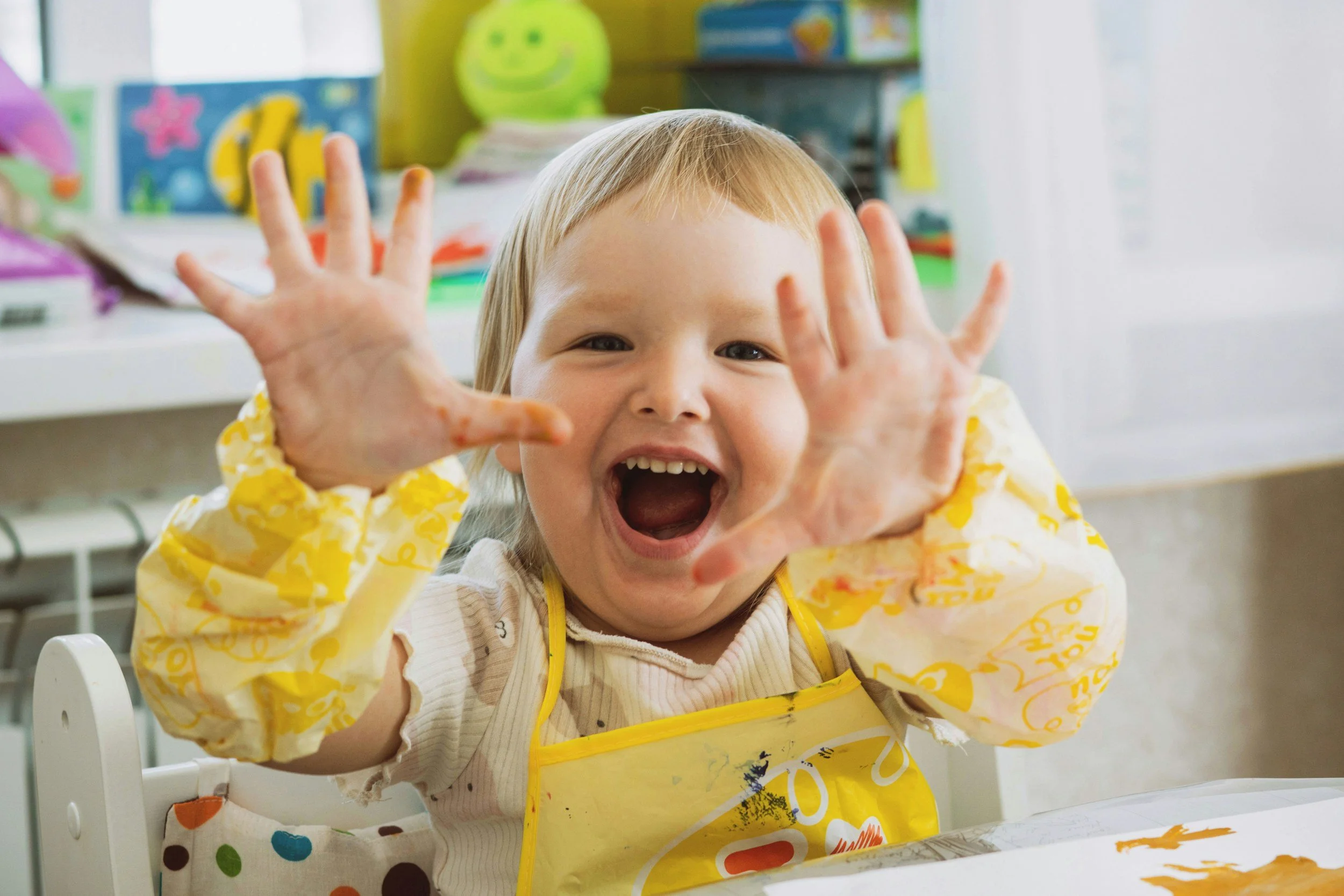 A joyful young girl with blonde hair wearing a yellow apron sits at a table with painted hands, smiling and reaching towards the camera in a colorful classroom or art room.