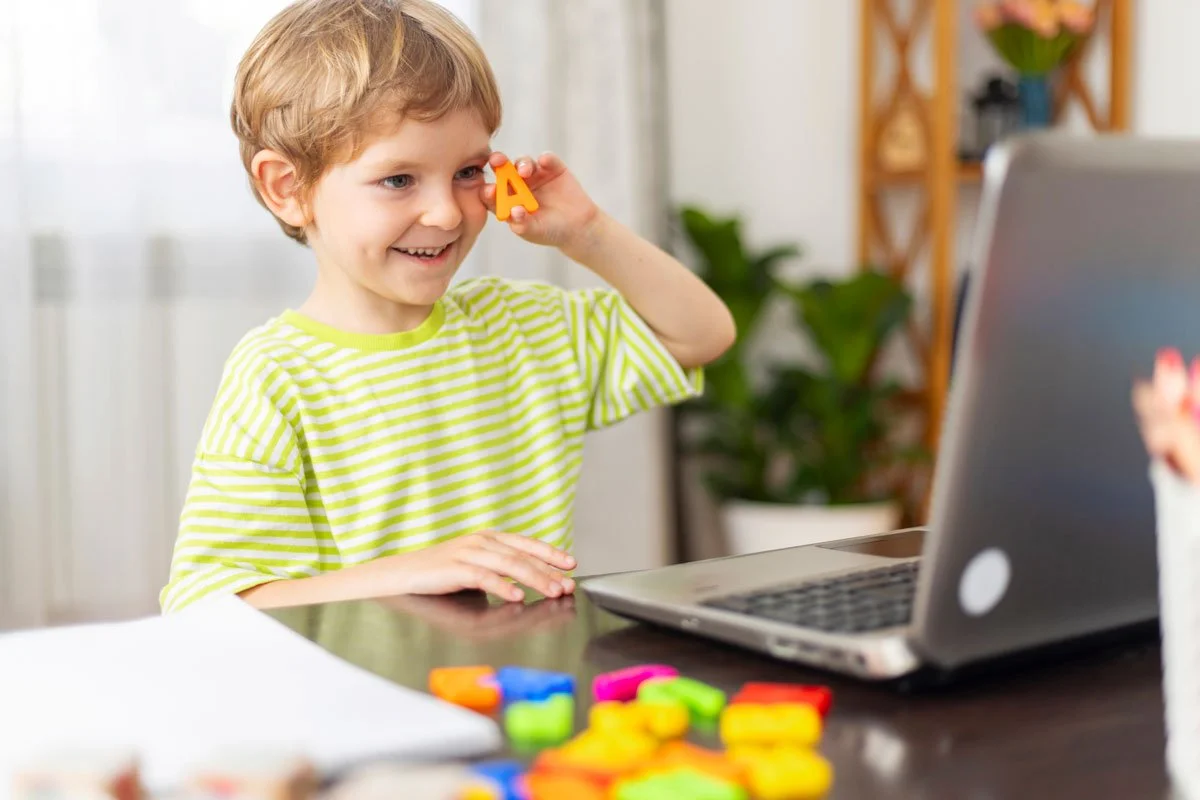 Young Caucasion boy in light green t-shirt, holding a letter A up near his face while doing Tele-Play Therapy in front of a laptop at home.