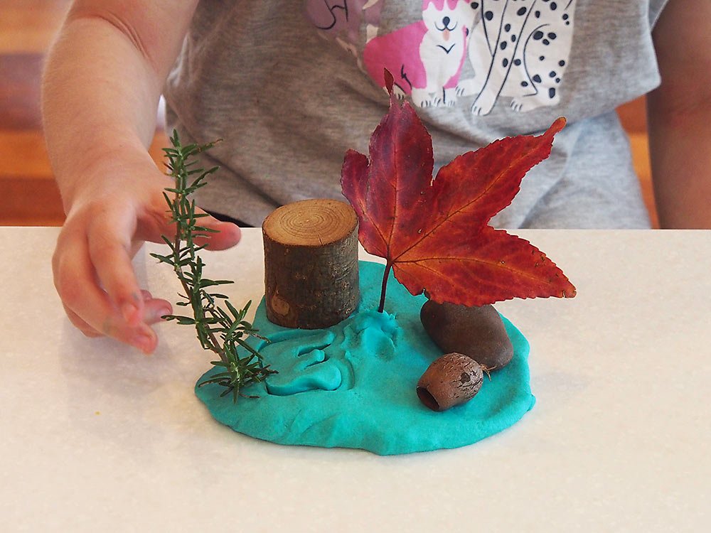 A child's hand arranging autumn-themed craft materials on turquoise clay, including a wooden log, pine cone, acorn, red maple leaf, and sprig of pine.