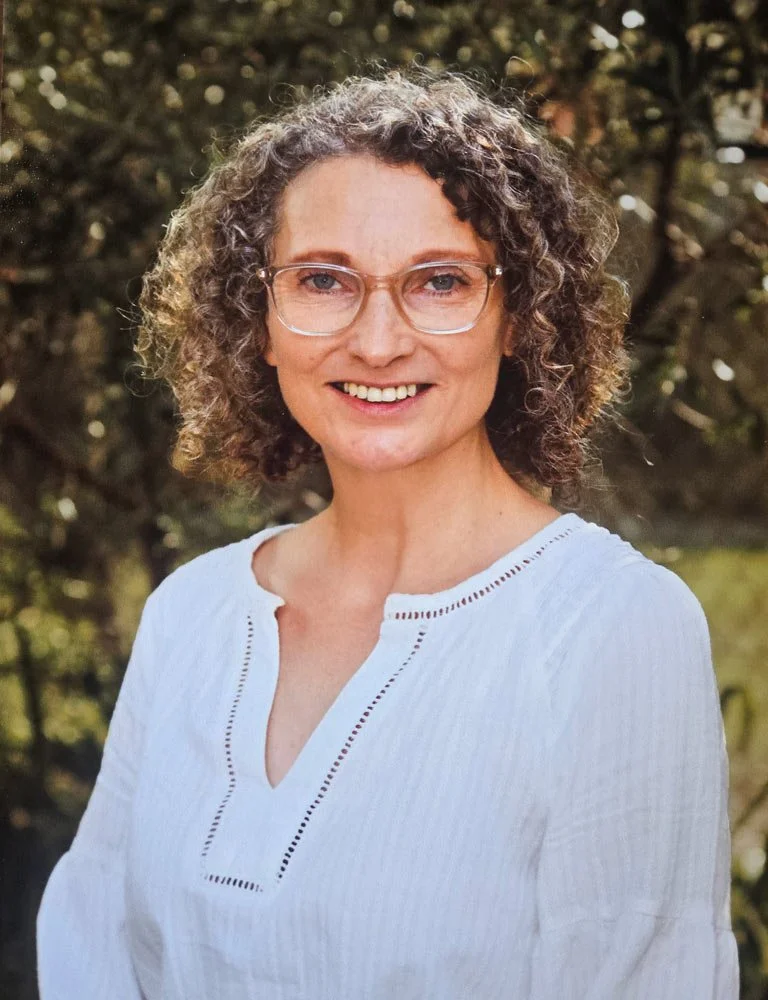 A woman with curly gray hair and glasses smiling outdoors.