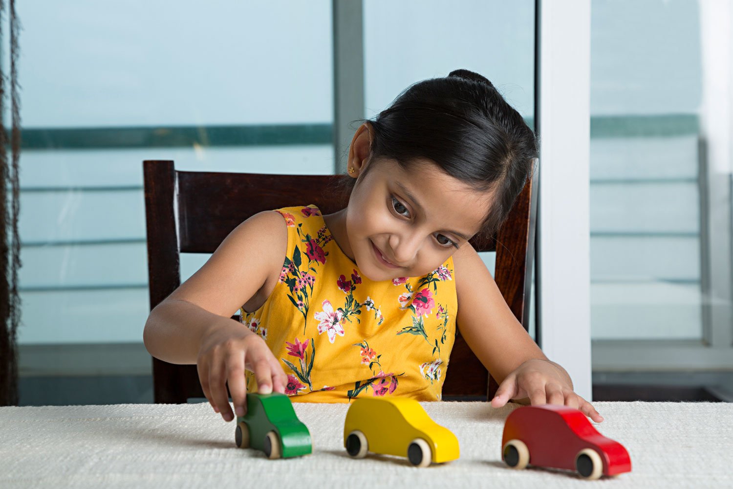 A young girl in a yellow floral dress plays with colorful wooden toy cars at a table in a bright room.