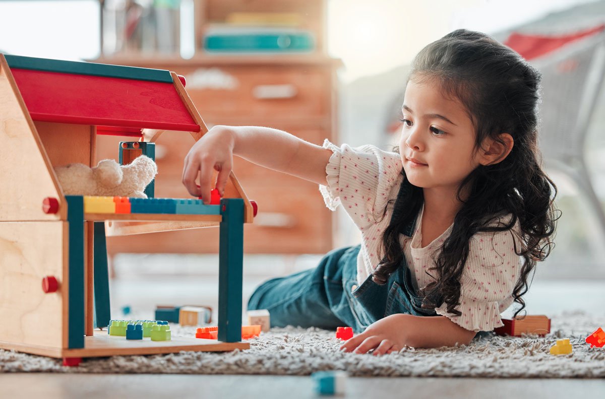 Young girl playing with colorful building blocks and a stuffed animal on a carpeted floor in a cozy room.