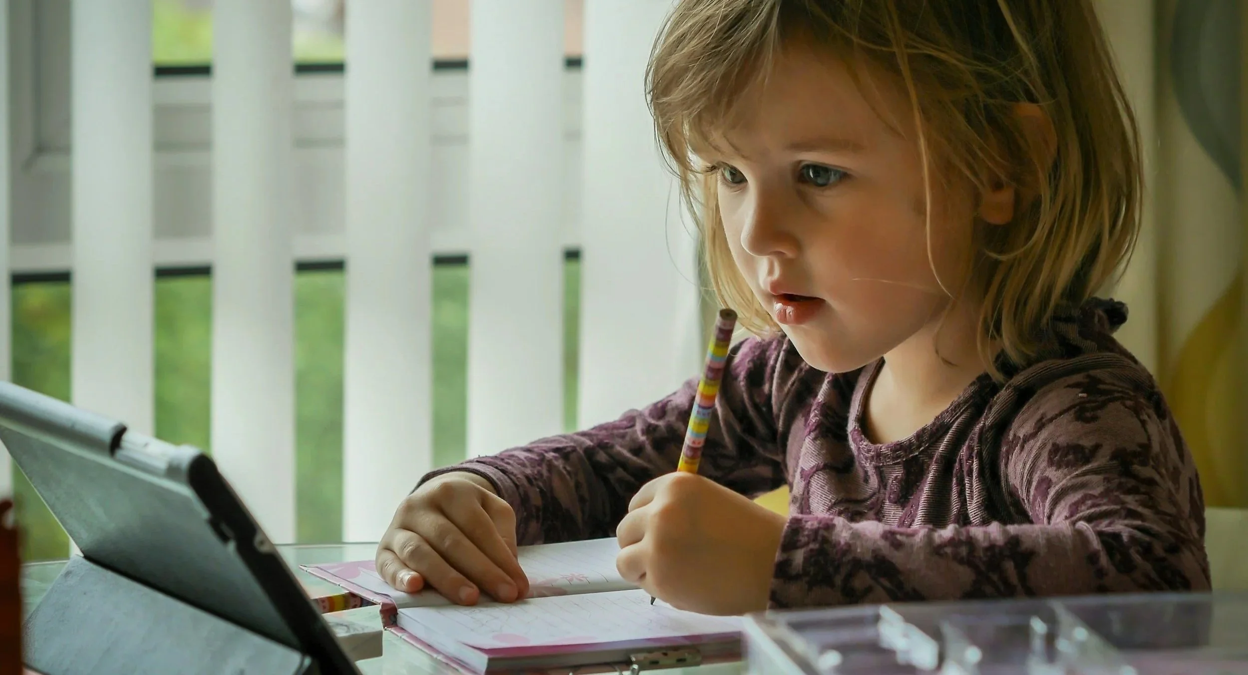 A young girl with shoulder-length blonde hair sitting at a table, writing in a notebook with a colorful striped pencil, while looking at a tablet device.