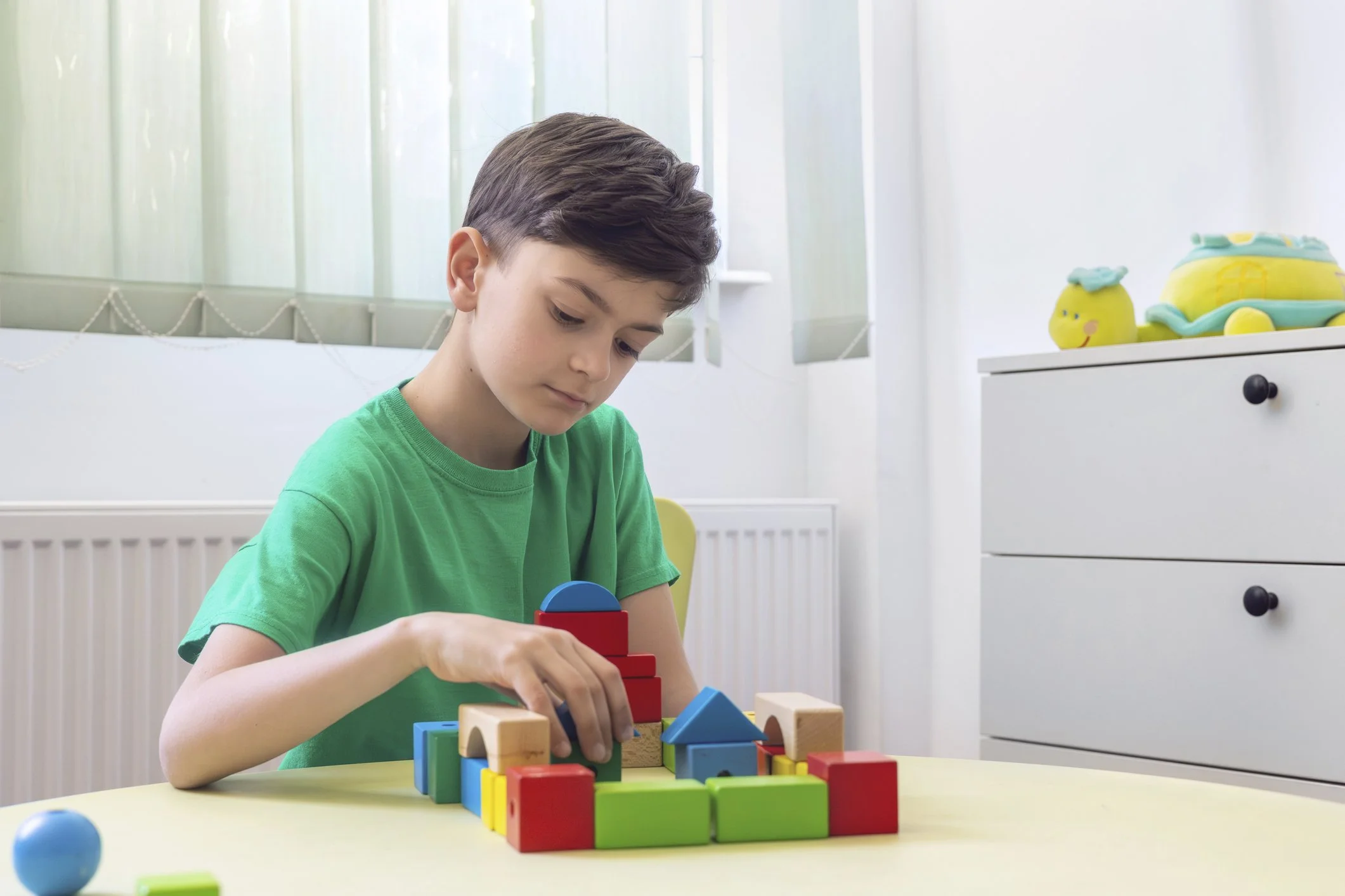 A young boy in a green T-shirt playing with colorful wooden building blocks on a yellow table in a bright room with a window and a toy turtle on a white dresser.