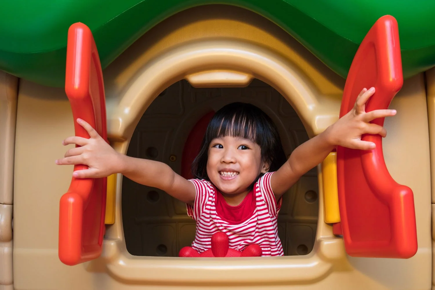 Smiling young Chinese girl looking out of the window of a cubby house with out-stretched arms .