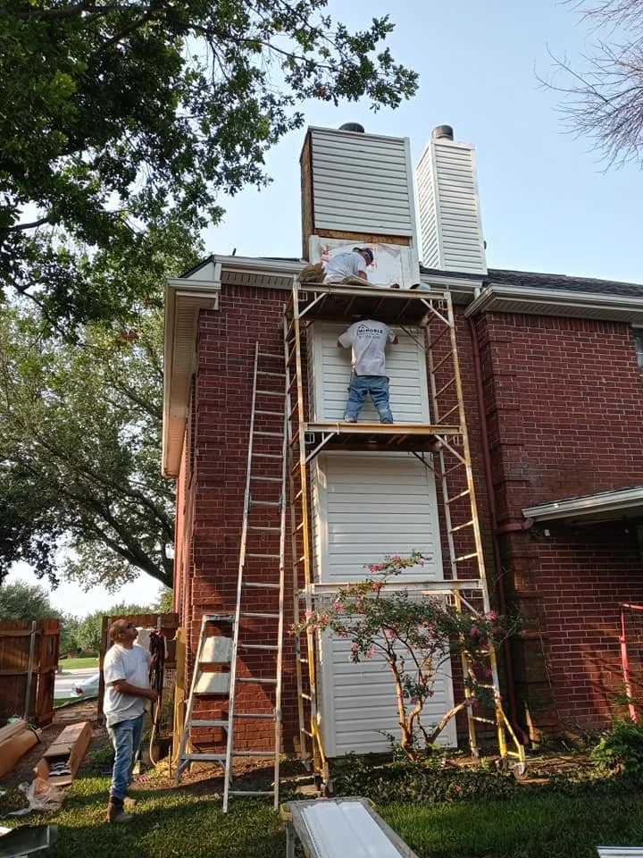 Workers installing or repairing siding on a two-story brick house. One worker is on scaffolding, while another is on the roof. A woman stands on the ground nearby, holding a tool or material. There is a ladder leaning against the house and trees in t